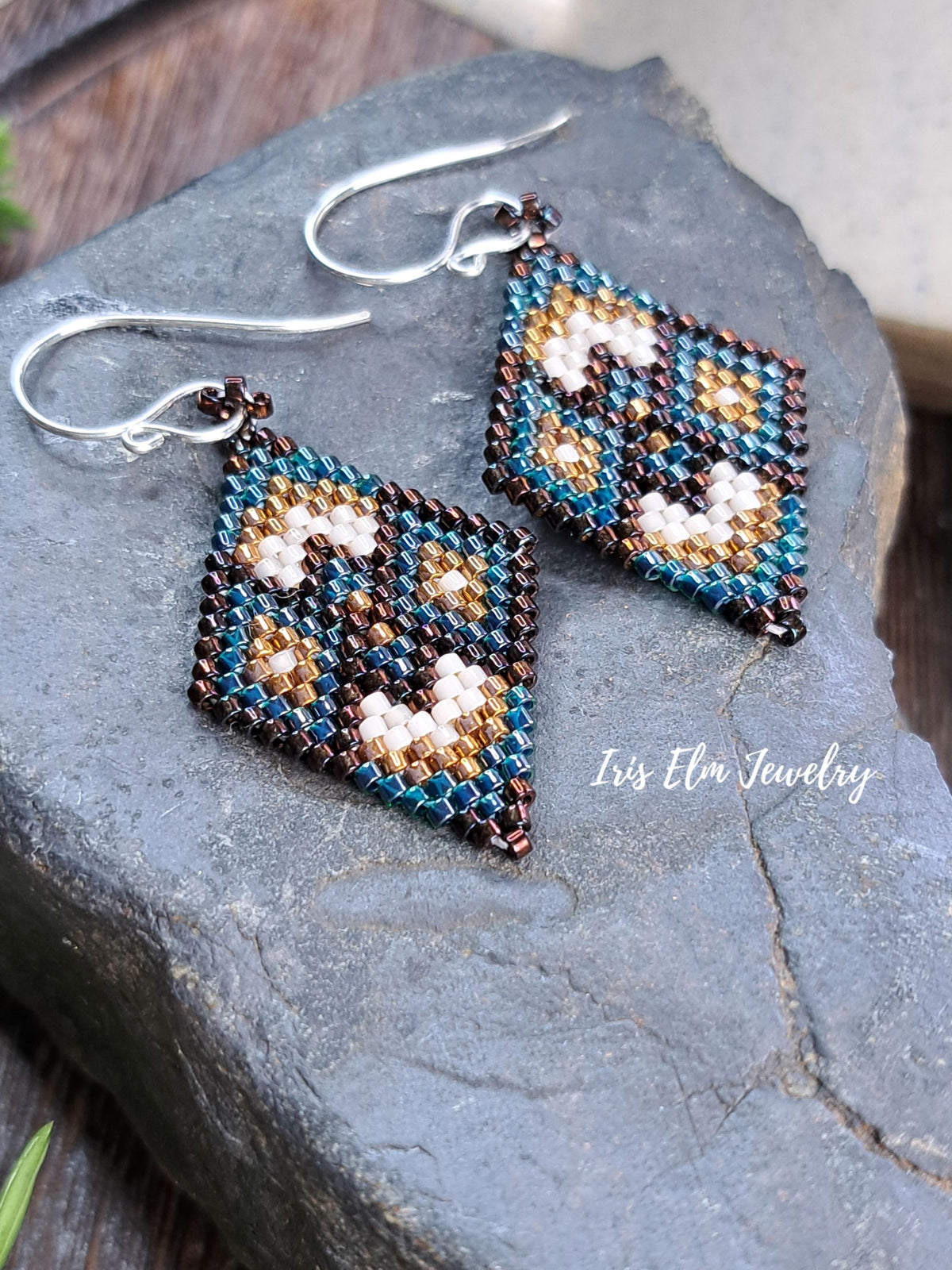 A pair of handmade beaded dangle earrings with silver fishhook wires, displayed on a dark gray slate stone. The earrings feature a geometric pattern in teal, brown, cream.