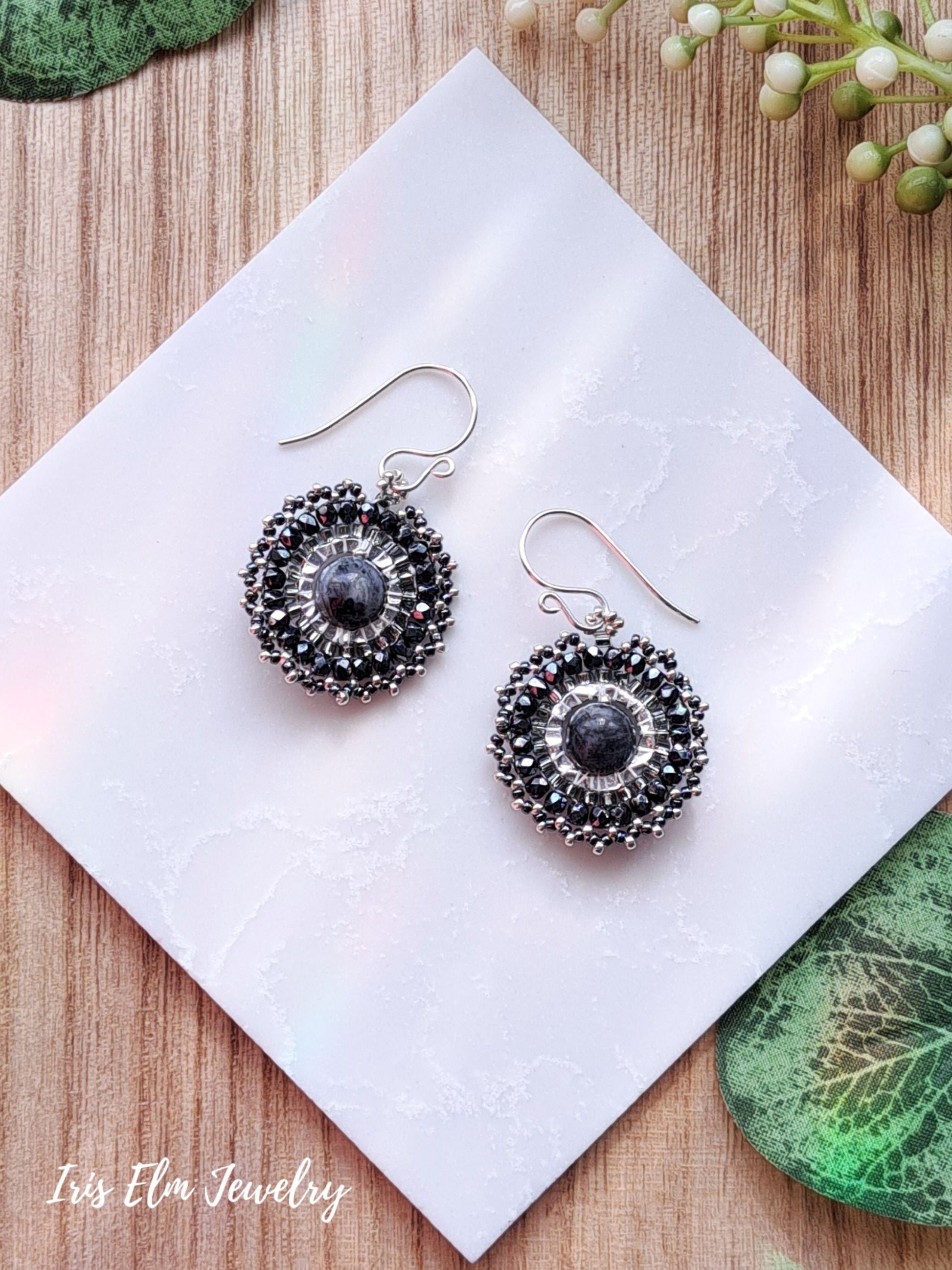 Pair of silver beadwork dark grey larvikite hoop earrings with black and grey stones on a white tile, surrounded by green leaves.
