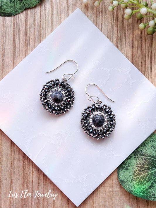 Pair of silver beadwork dark grey larvikite hoop earrings with black and grey stones on a white tile, surrounded by green leaves.
