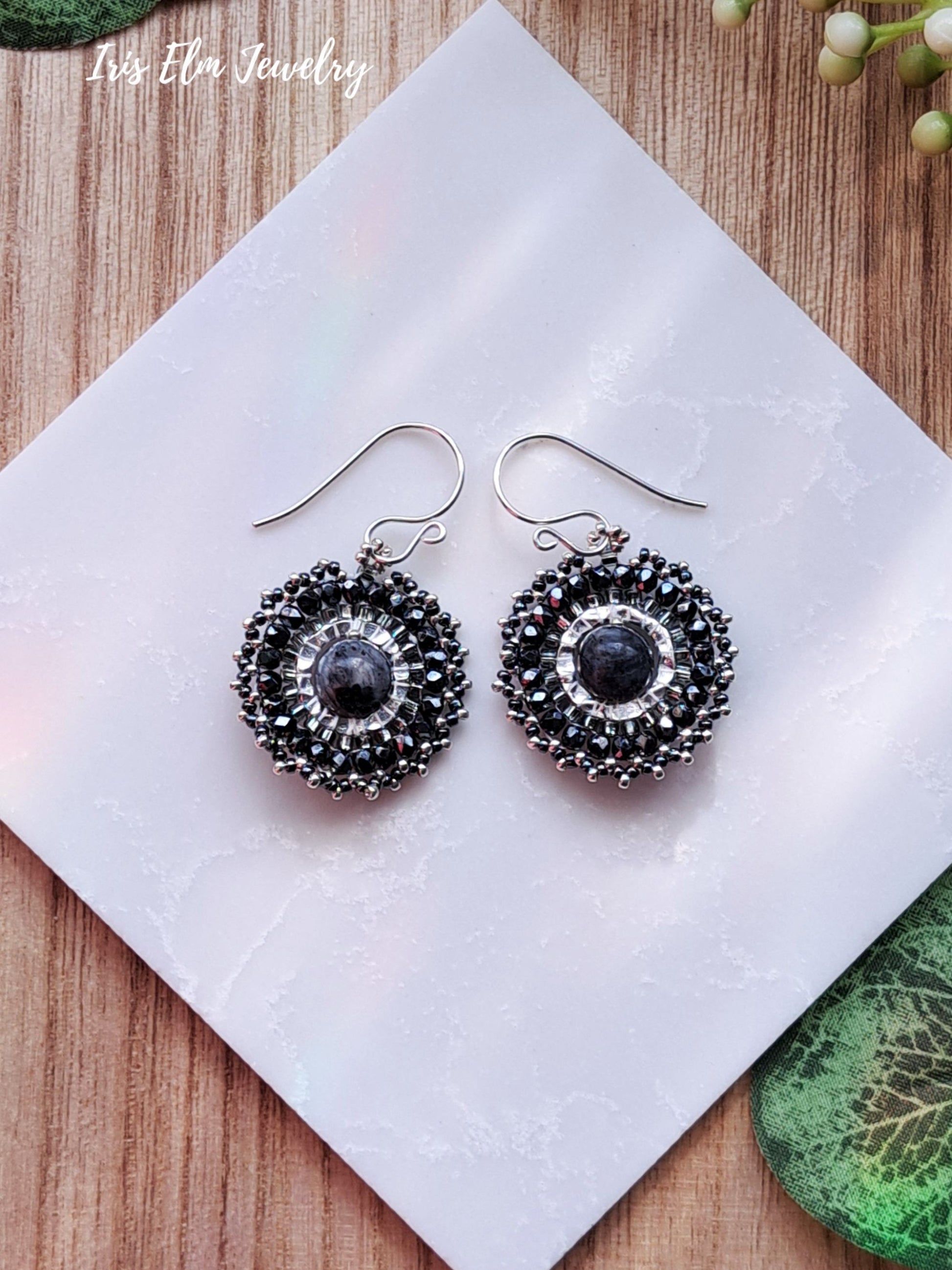 Pair of silver glass beadwork woven hoop earrings with black larvikite stones on a white napkin with 'Iris Elm Jewelry' branding.