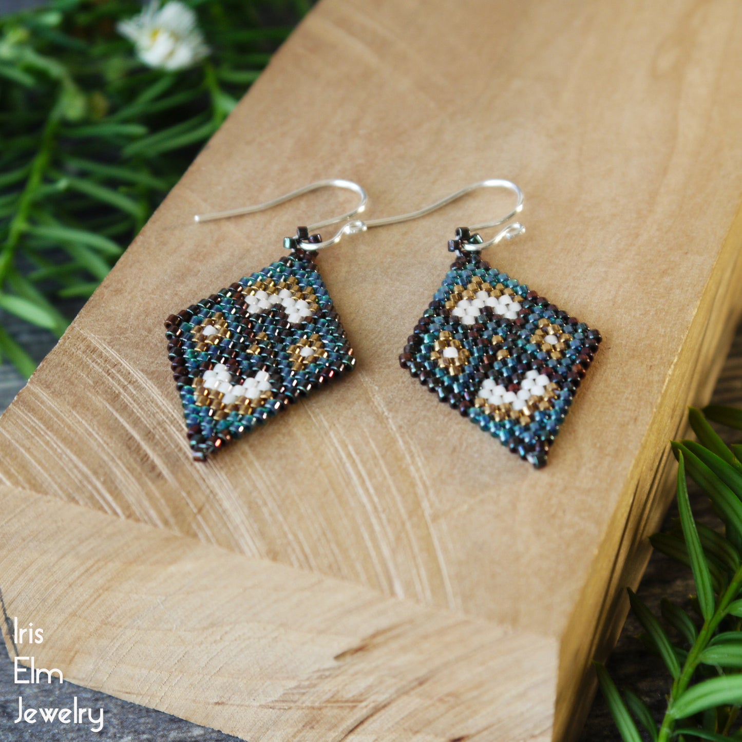 Pair of beaded forest green and bronze glass earrings on a wooden surface with greenery in the background
