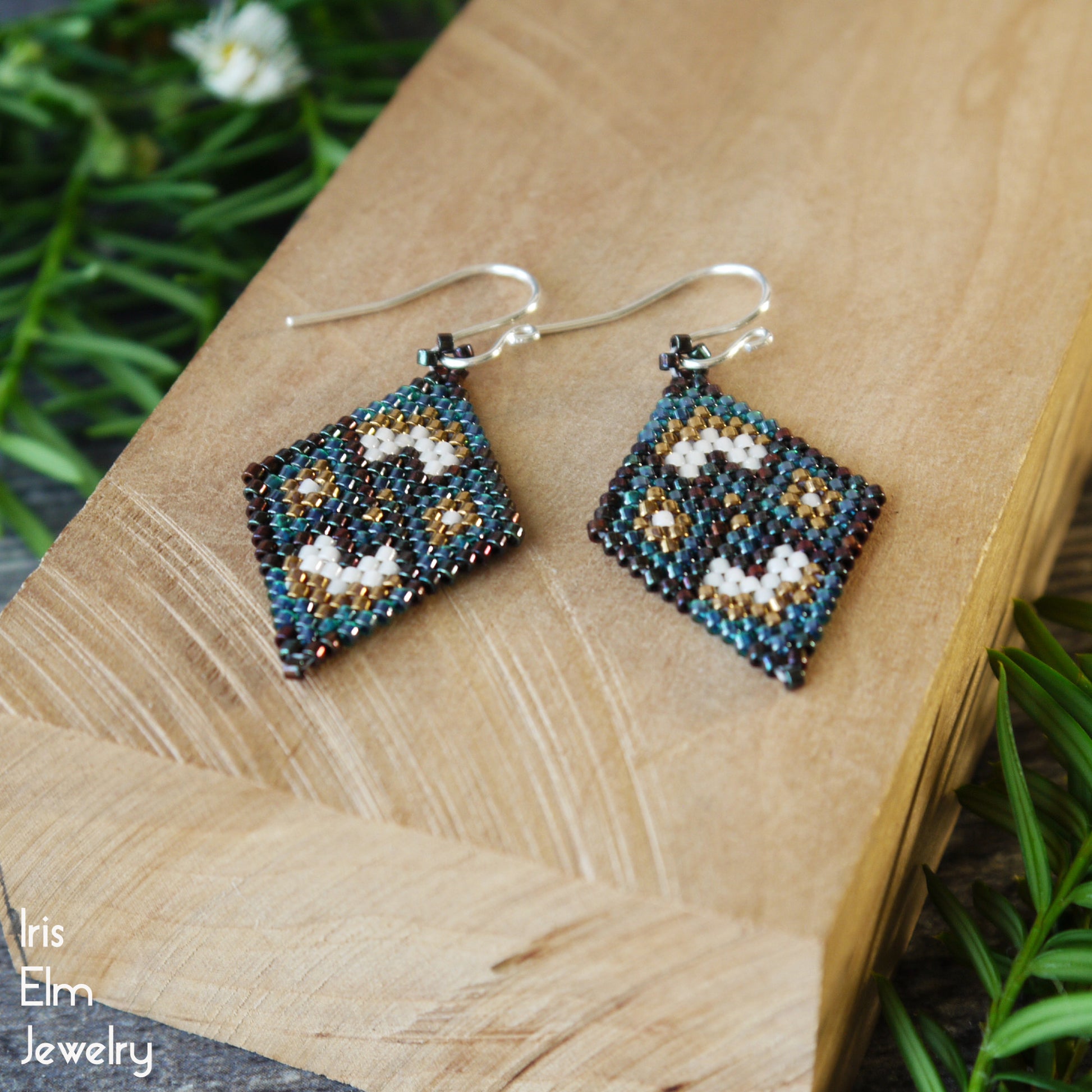 Pair of beaded forest green and bronze glass earrings on a wooden surface with greenery in the background