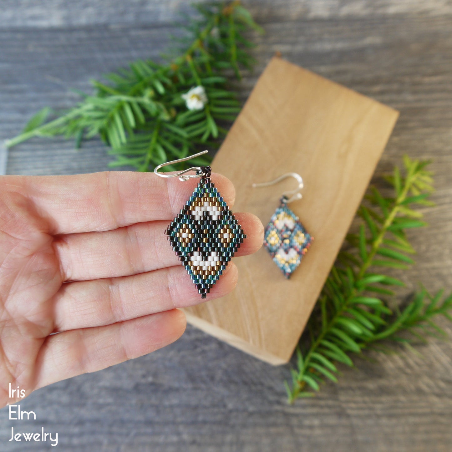 Hand holding a earth tone beaded diamond-shaped earring with another similar earring on a wooden block, against a natural background.