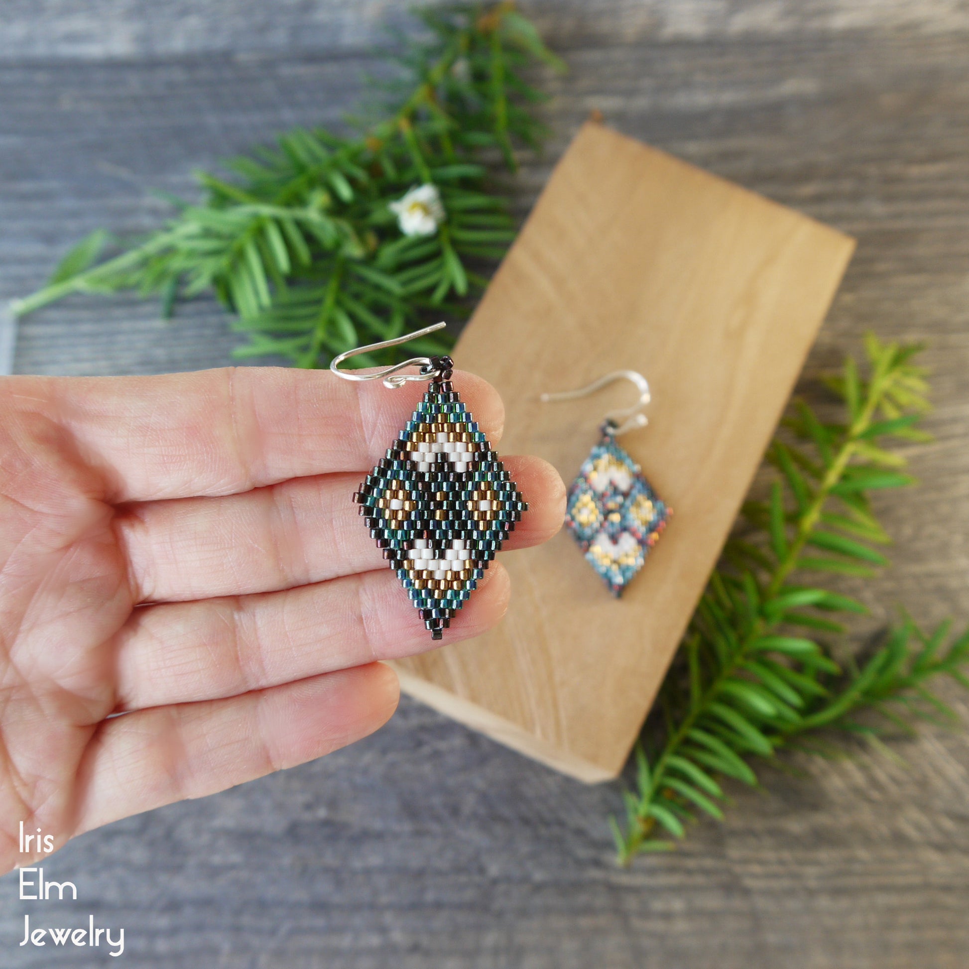 Hand holding a earth tone beaded diamond-shaped earring with another similar earring on a wooden block, against a natural background.