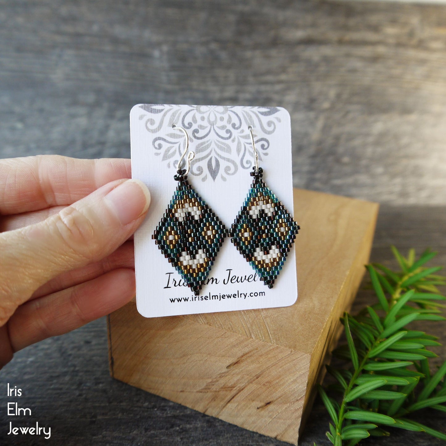 Hand holding a pair of beaded earrings on a branded card with a wooden block and plant in the background.