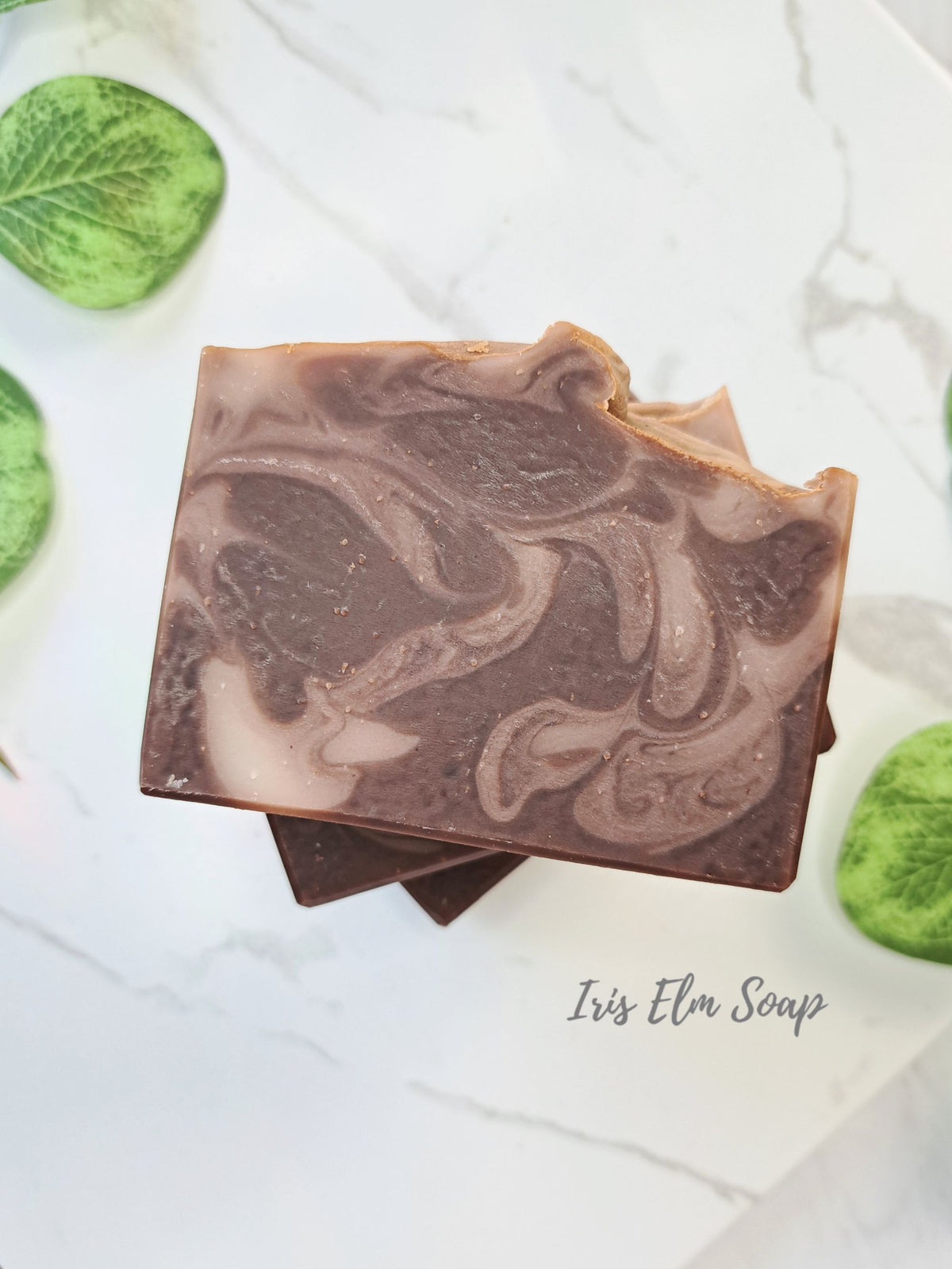 A stack of artisanal marbled soap bars in shades of brown and beige, styled on a white marble surface with green leaves. The text 'Iris Elm Soap' is visible in the bottom right corner.
