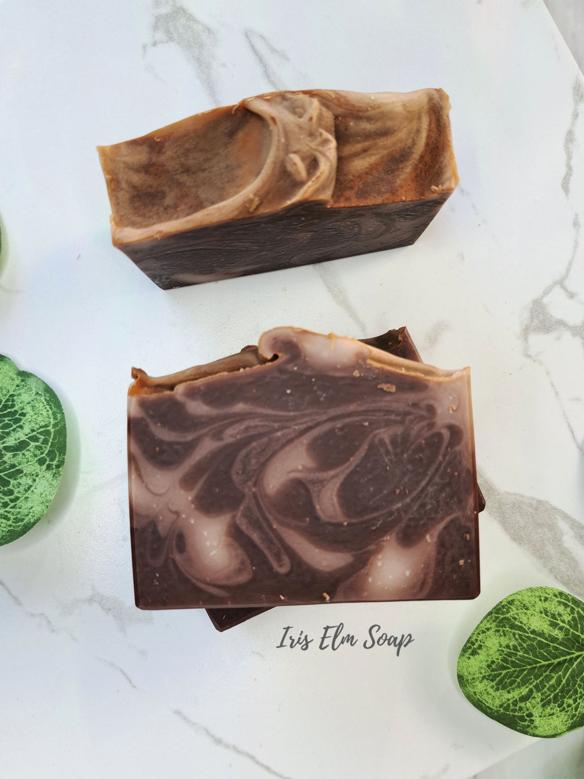 Two vanilla oatmeal artisanal marbled soap bars displayed on a white marble surface with decorative green leaves. The top bar is light brown and dark brown, and the bottom bar is a rich chocolate brown with lighter cream swirls.