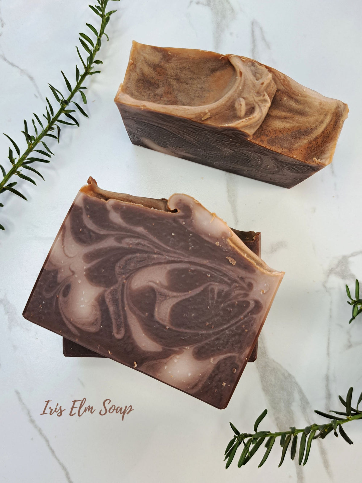 Two artisanal handcrafted soap bars with marbled brown and beige swirls, displayed on a white marble surface with green rosemary sprigs. The brand name 'Iris Elm Soap' is visible in the bottom left corner.