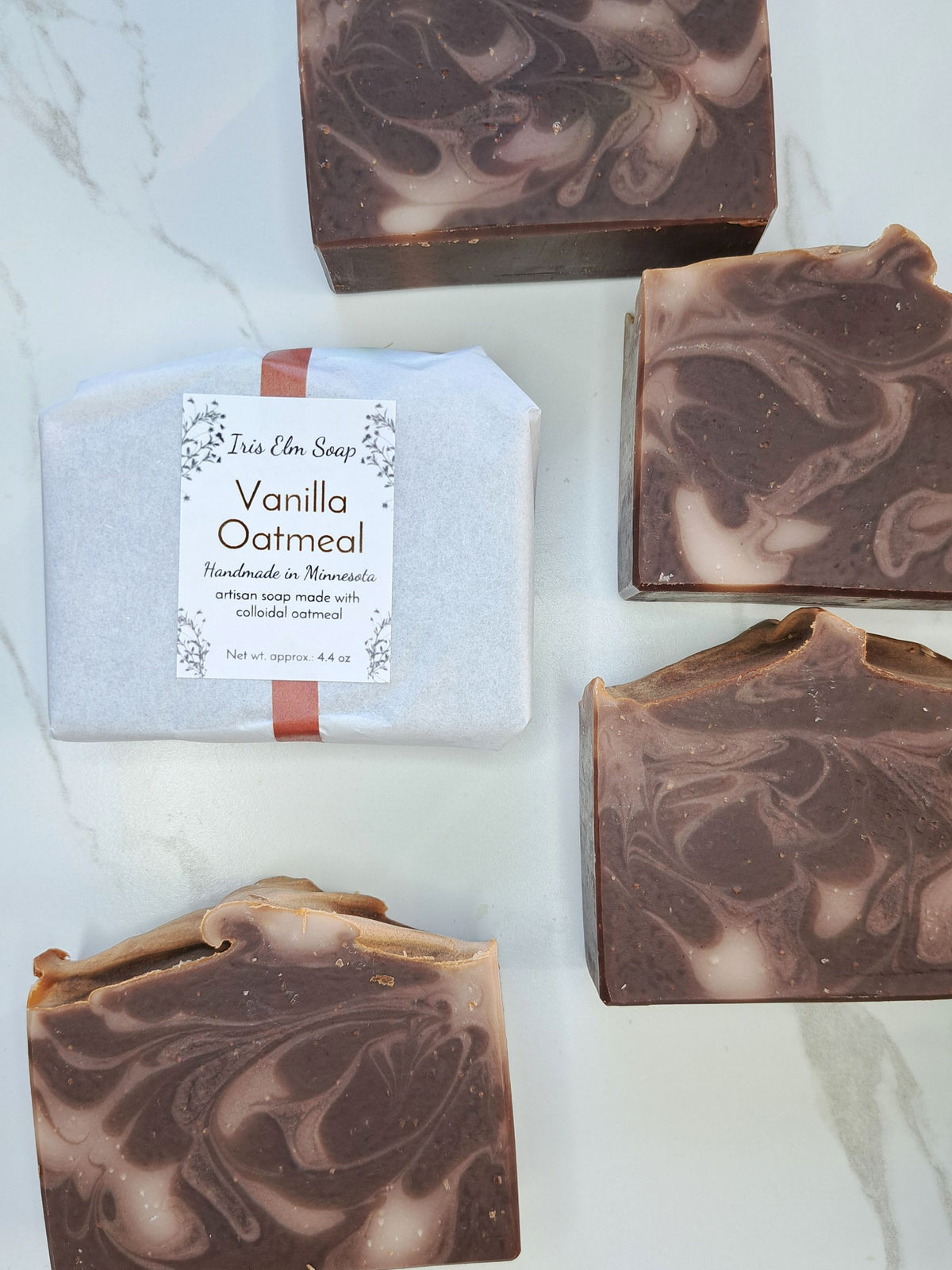 Four rectangular artisan soap bars with a brown and cream marbled swirl pattern are arranged on a white marble surface, alongside a wrapped bar labeled 'Vanilla Oatmeal' by Iris Elm Soap.