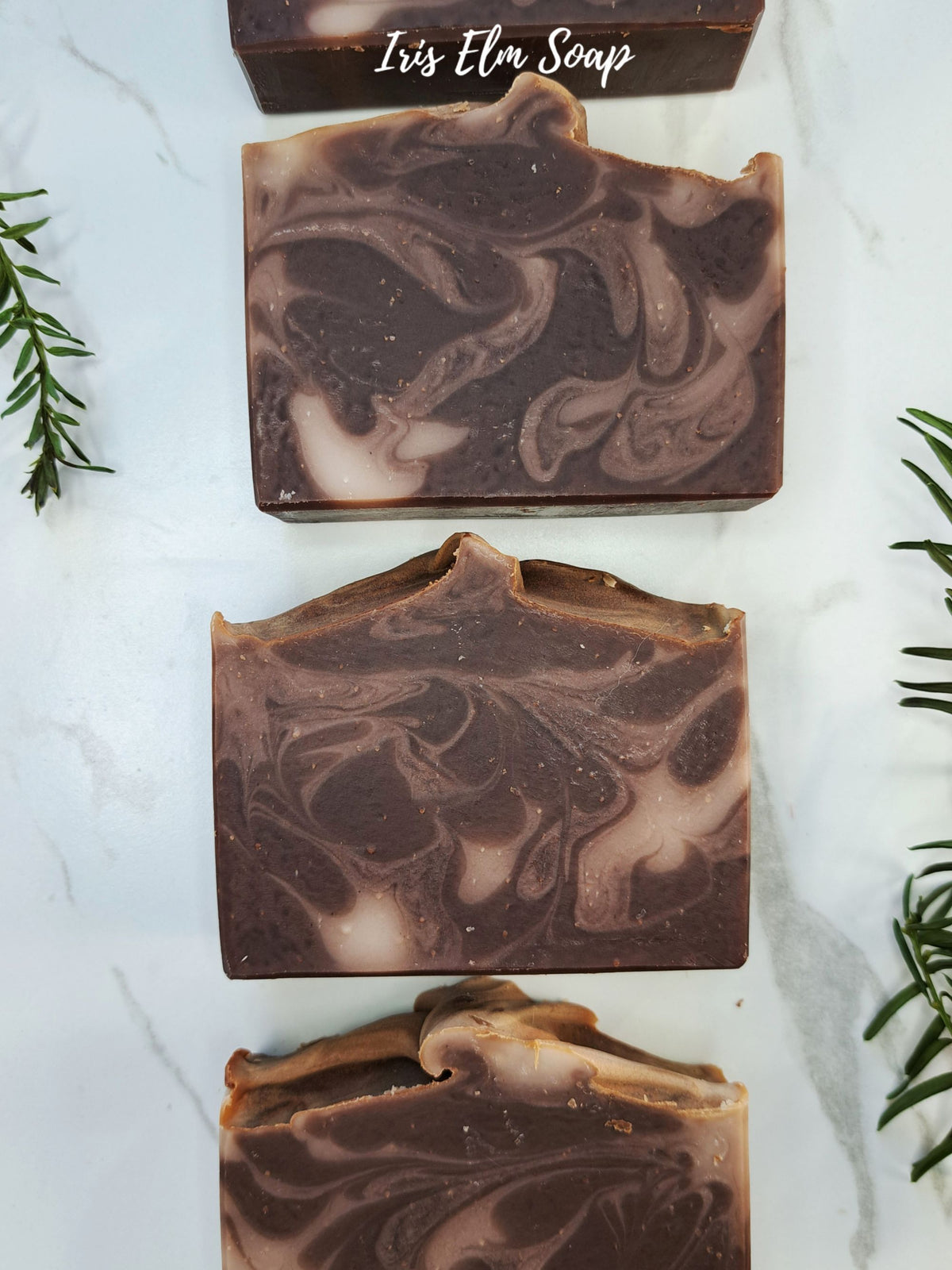 Three handcrafted rectangular bars of vanilla oatmeal soap with a dark brown and light beige marbled swirl pattern, arranged vertically on a white marble surface with rosemary sprigs and a brand label 'Iris Elm Soap' at the top.