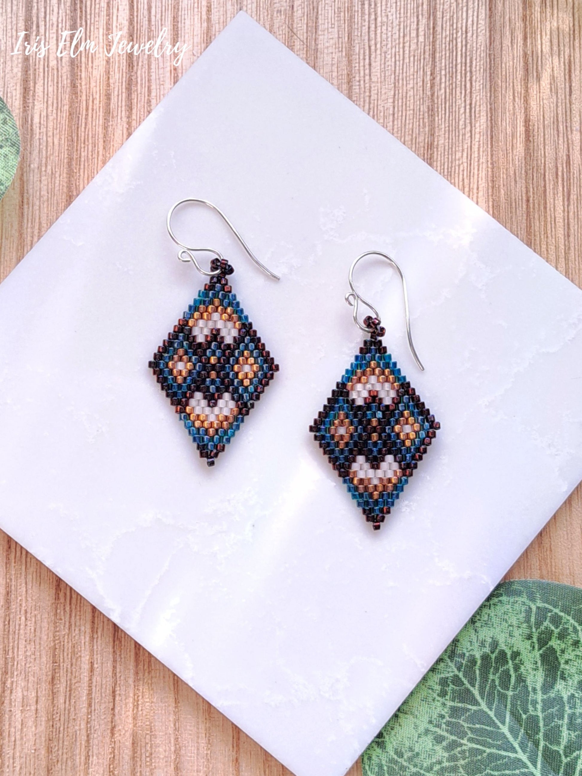 Pair of beaded brown and green geometric diamond seed bead earrings on a white tile with a wooden background