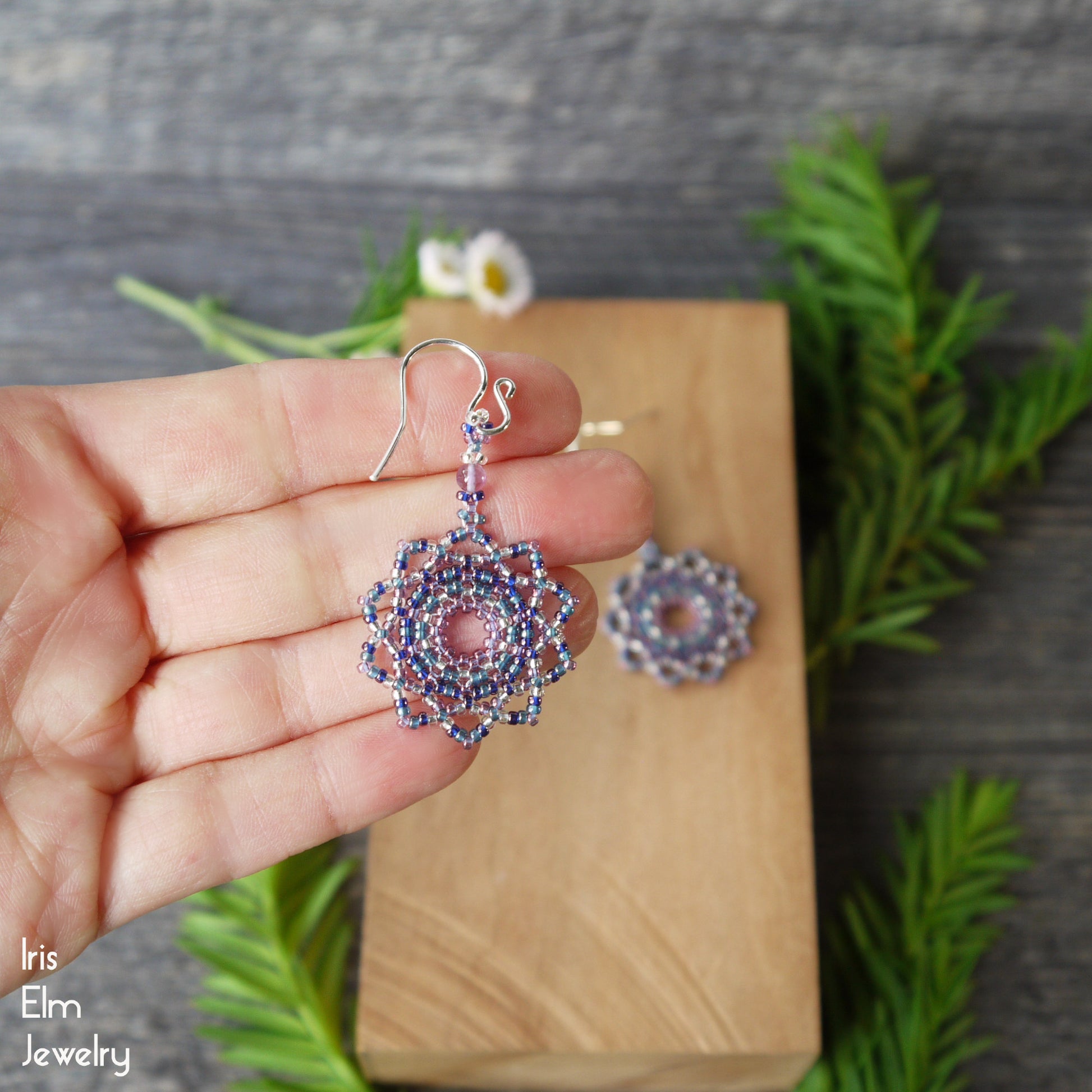Hand holding a pair of beaded purple seed bead mandala amethyst earrings with sterling silver ear wires above a wooden block and greenery in the background