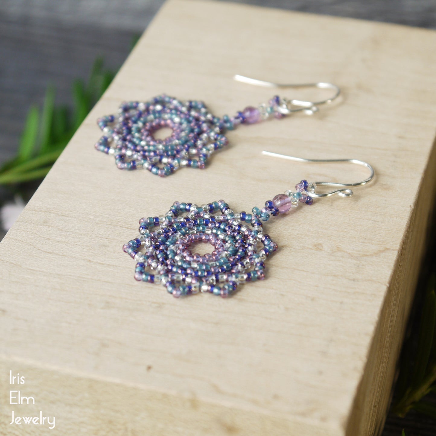 Beaded purple glass seed bead mandala earrings with amethyst gemstones on a wooden block with 'Iris Elm Jewelry' branding.