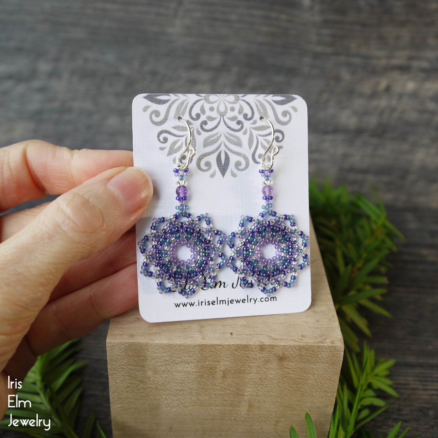Purple beaded earrings on a display card held by a hand with a wooden block and greenery background.