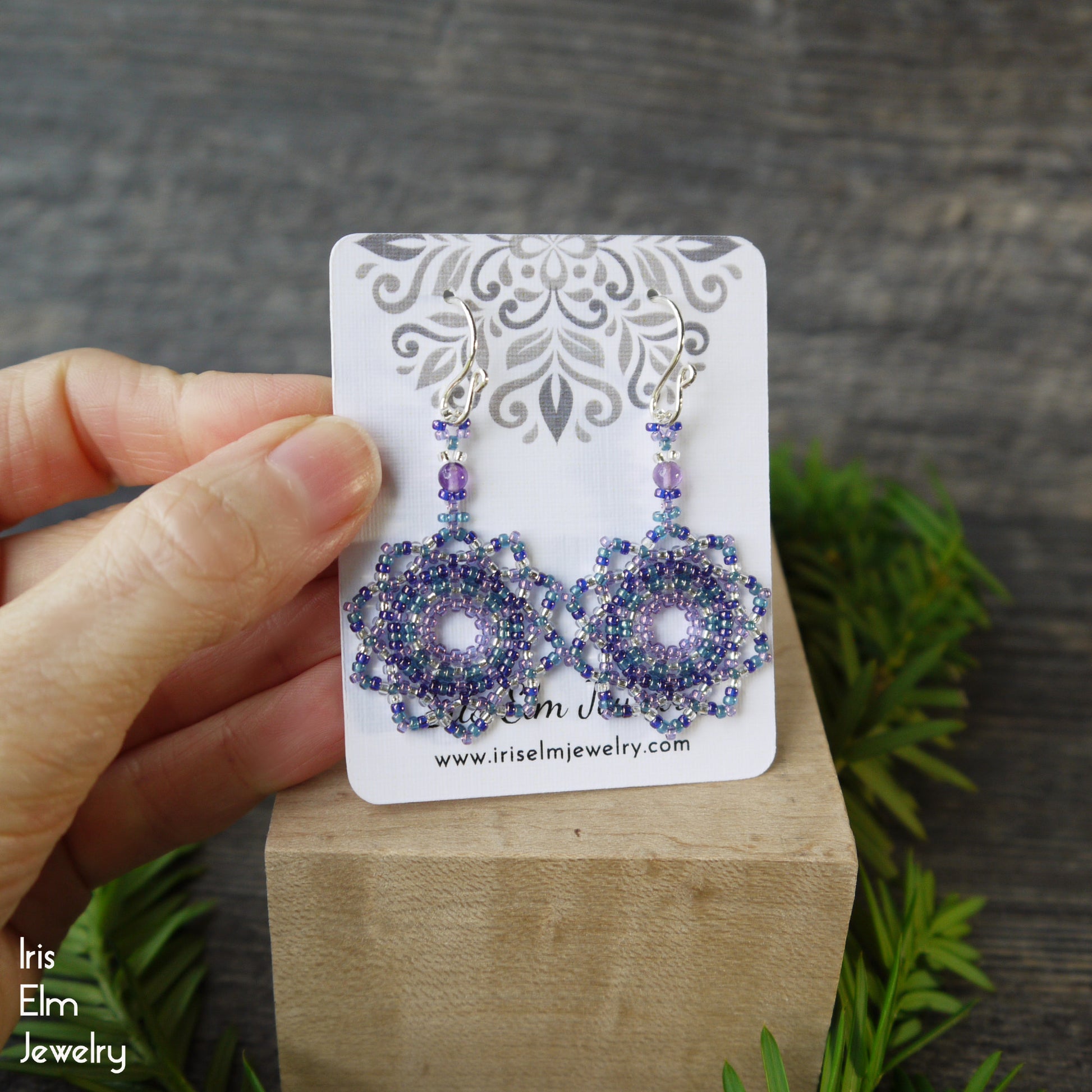 Purple beaded earrings on a display card held by a hand with a wooden block and greenery background.