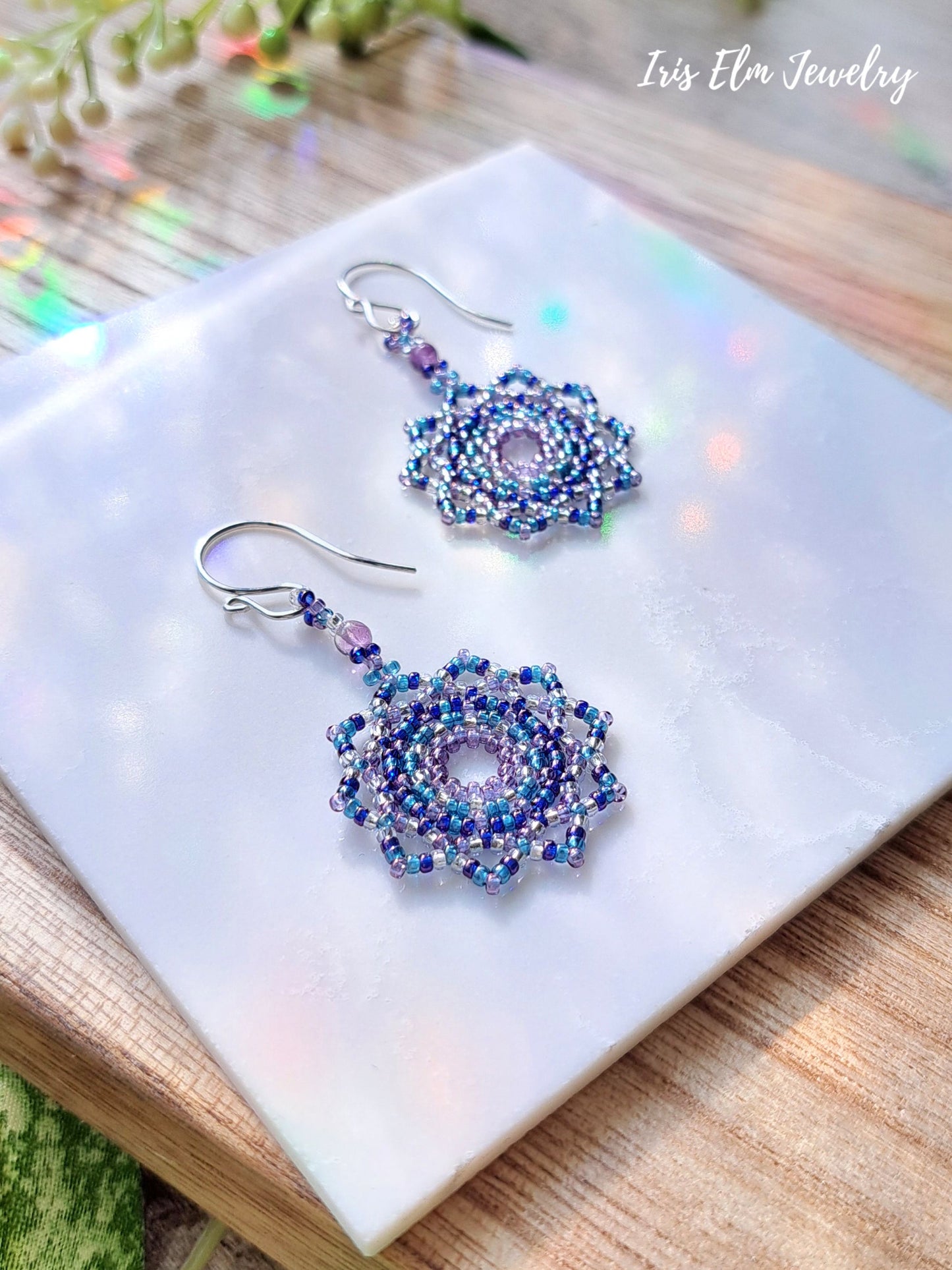 Pair of beaded purple mandala seed bead and amethyst earrings on a white marble surface with 'Iris Elm Jewelry' branding.