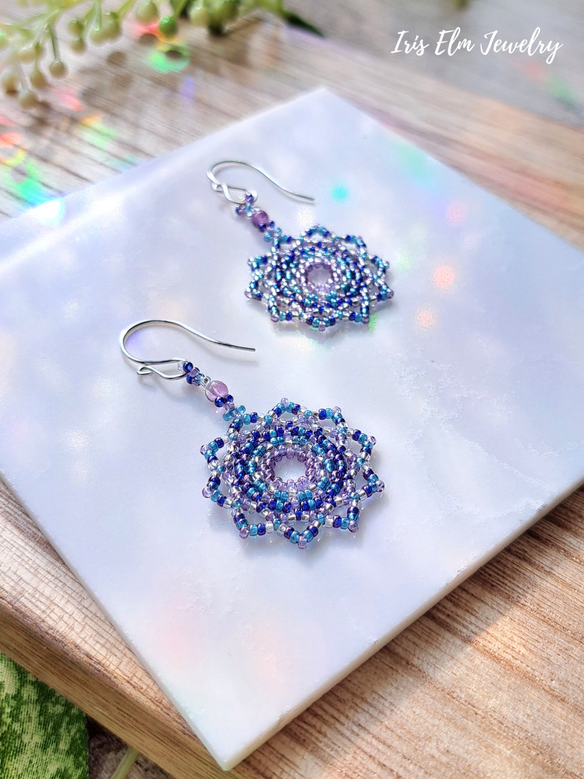 Pair of beaded purple mandala seed bead and amethyst earrings on a white marble surface with 'Iris Elm Jewelry' branding.