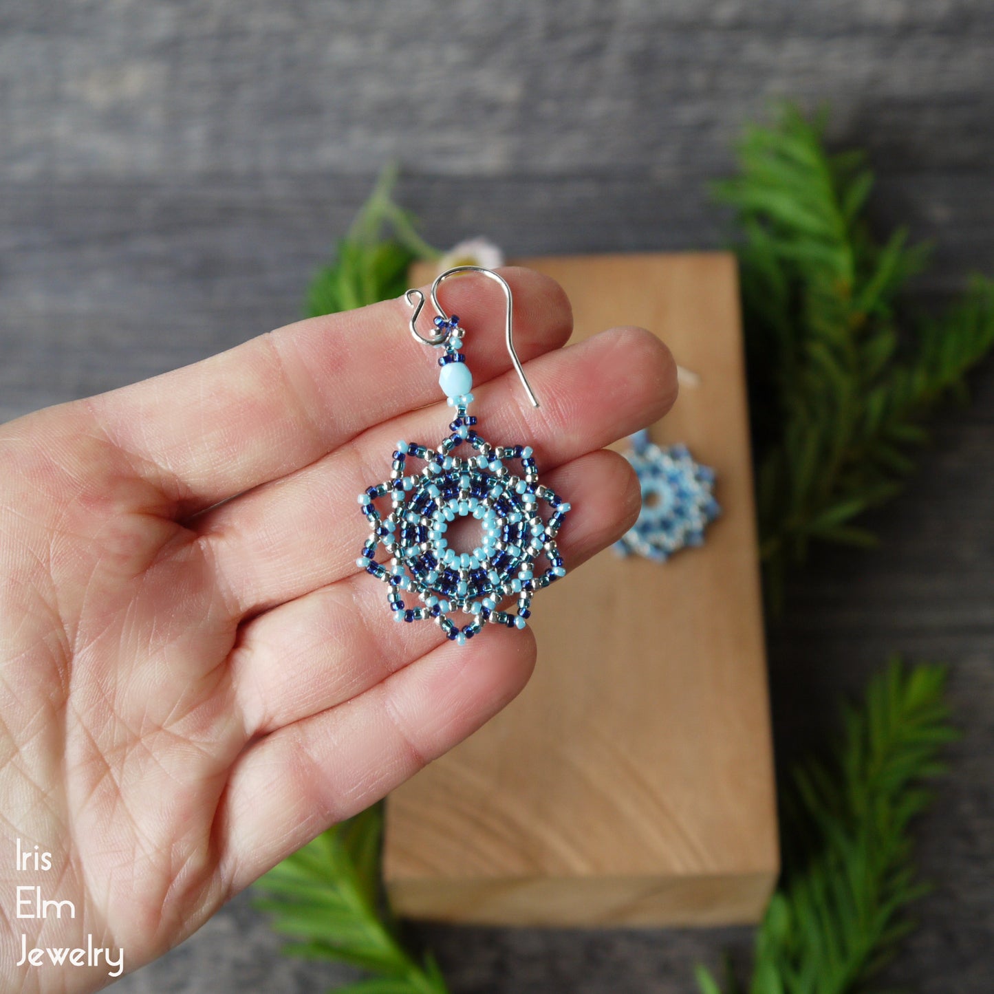 Hand holding a blue beaded woven mandala earring with sterling silver ear wire with a wooden box and greenery in the background 