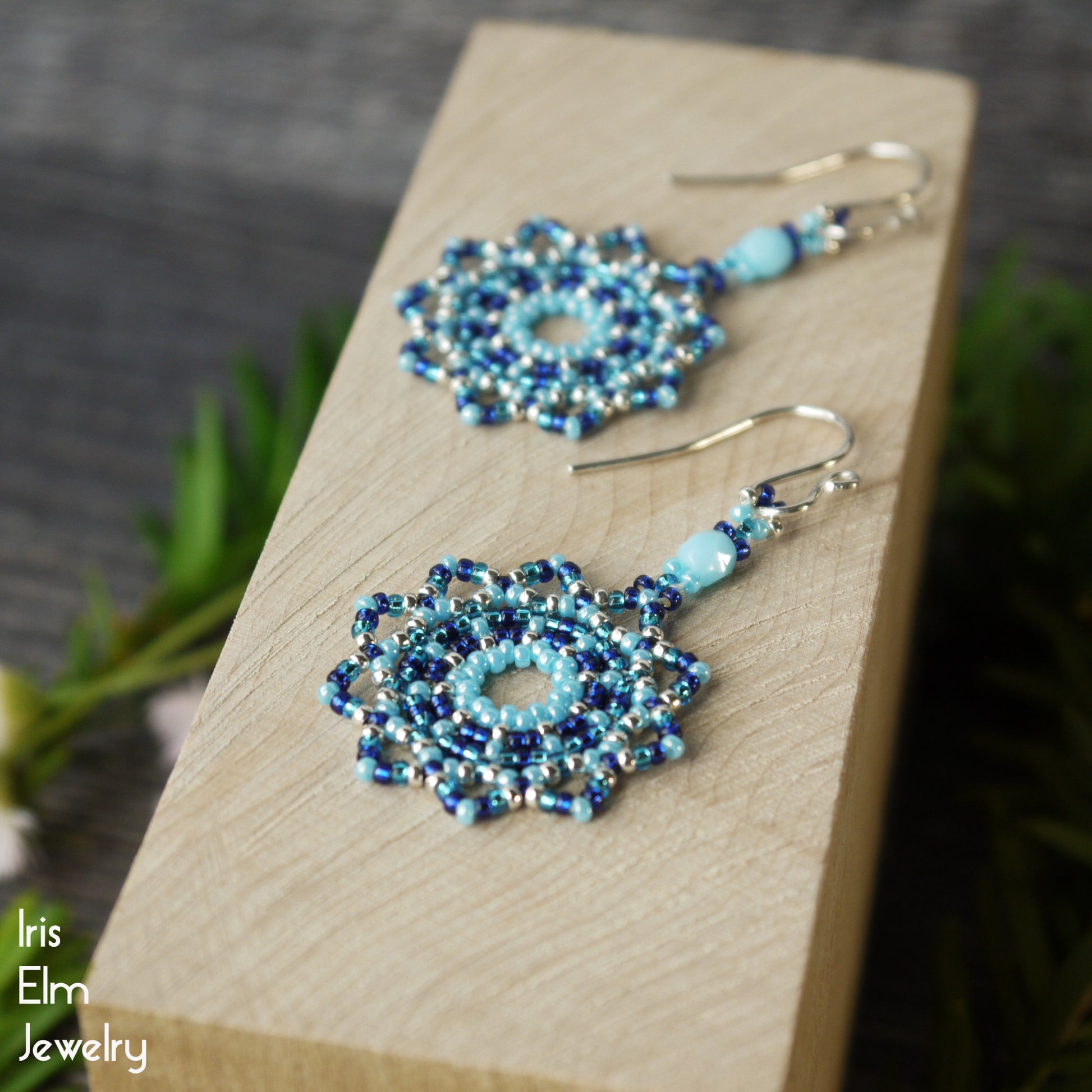 Pair of beaded blue seed bead mandala earrings on a wooden block with 'Iris Elm Jewelry' branding.