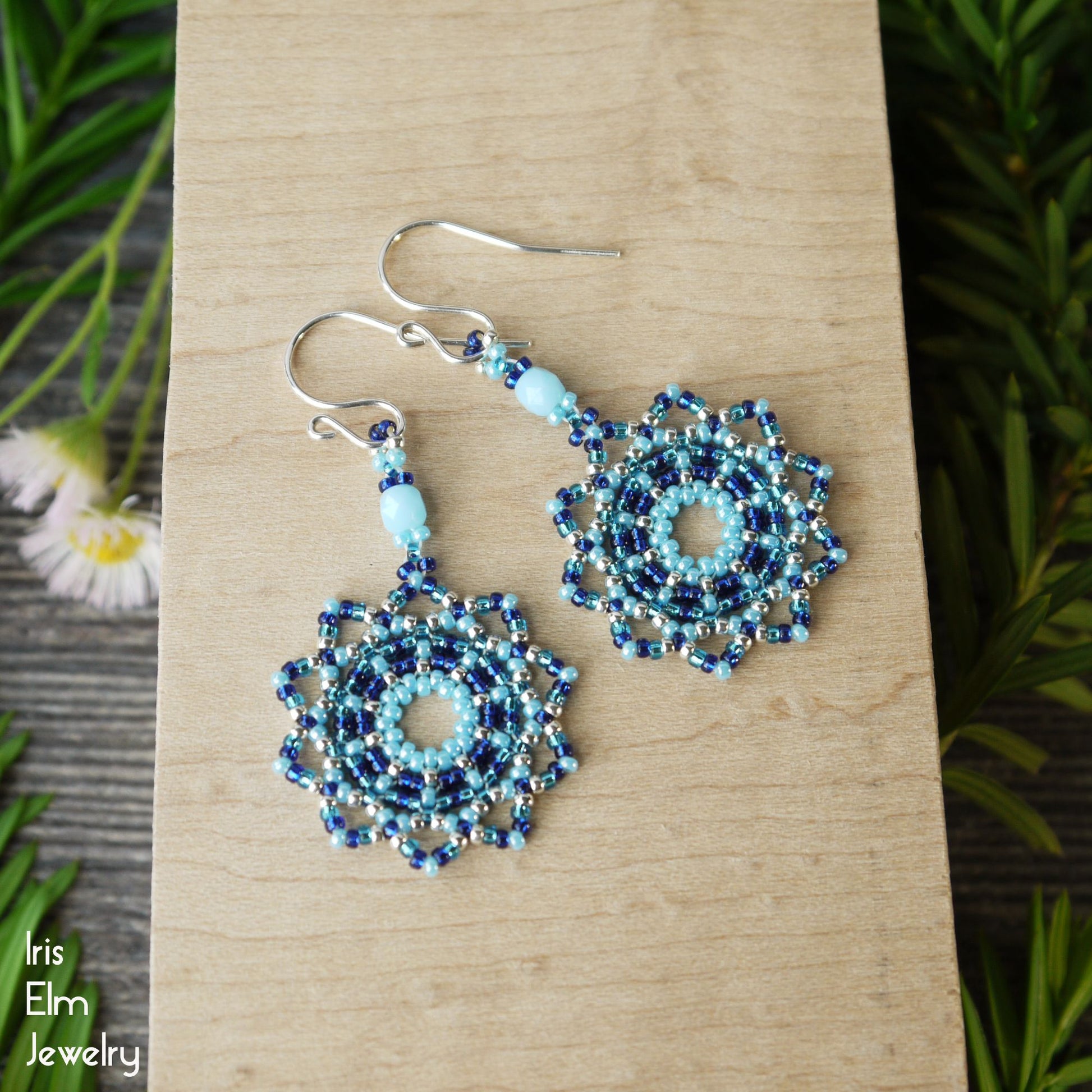 Pair of beaded seed bead woven mandala blue earrings on a wooden surface with greenery in the background, featuring the brand 'Iris Elm Jewelry'.