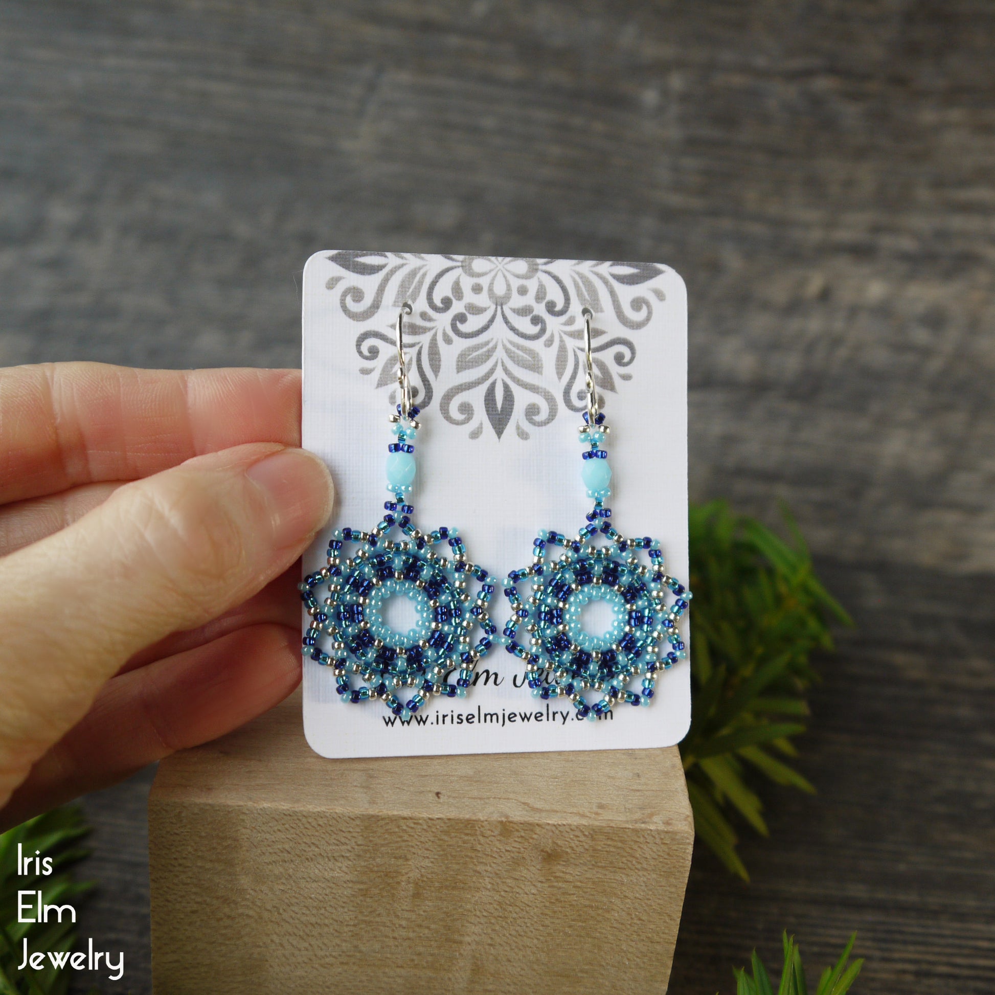 Blue beaded earrings on a card held by a hand with a wooden block and green leaves in the background.
