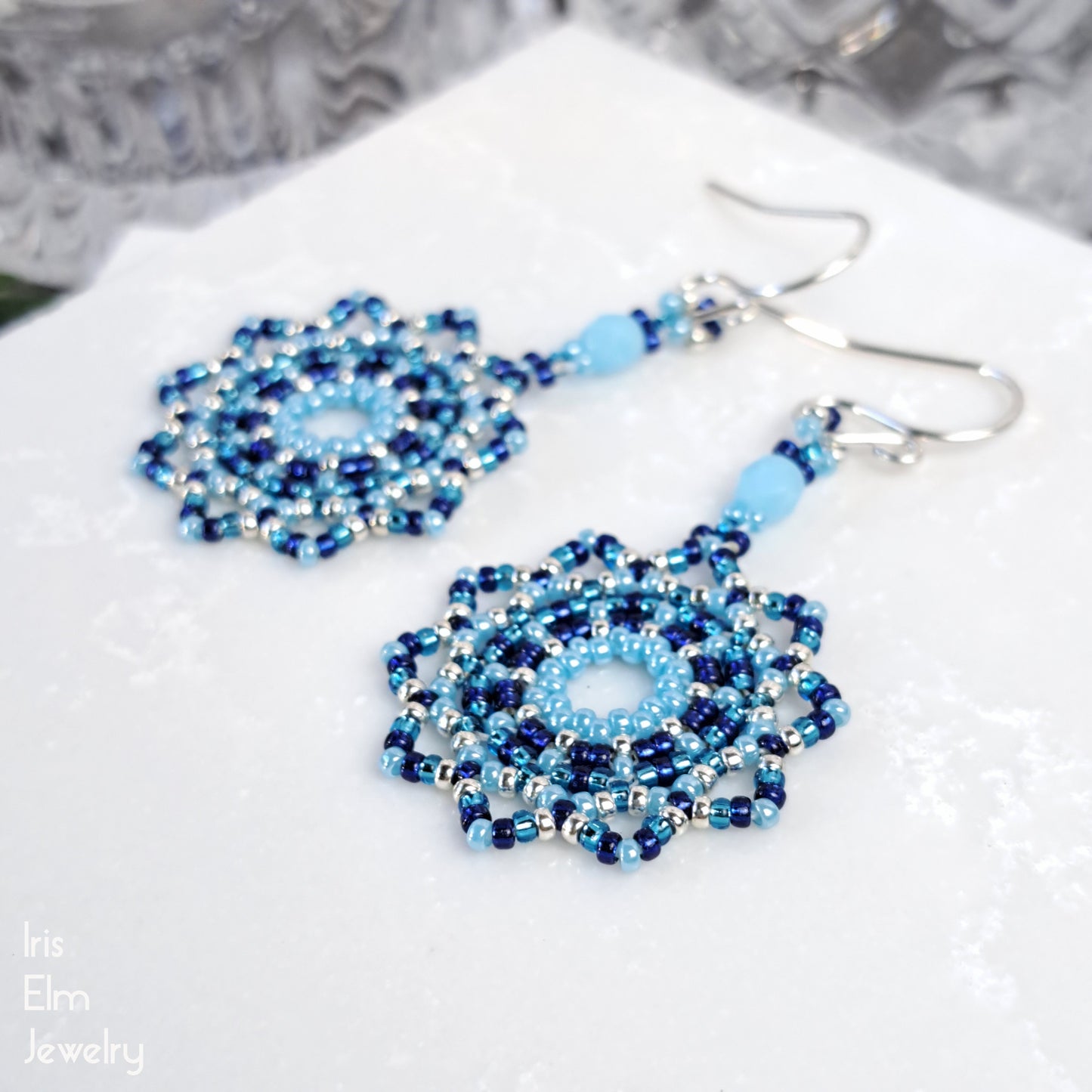 Blue beaded seed bead mandala earrings on a white surface with 'Iris Elm Jewelry' branding.