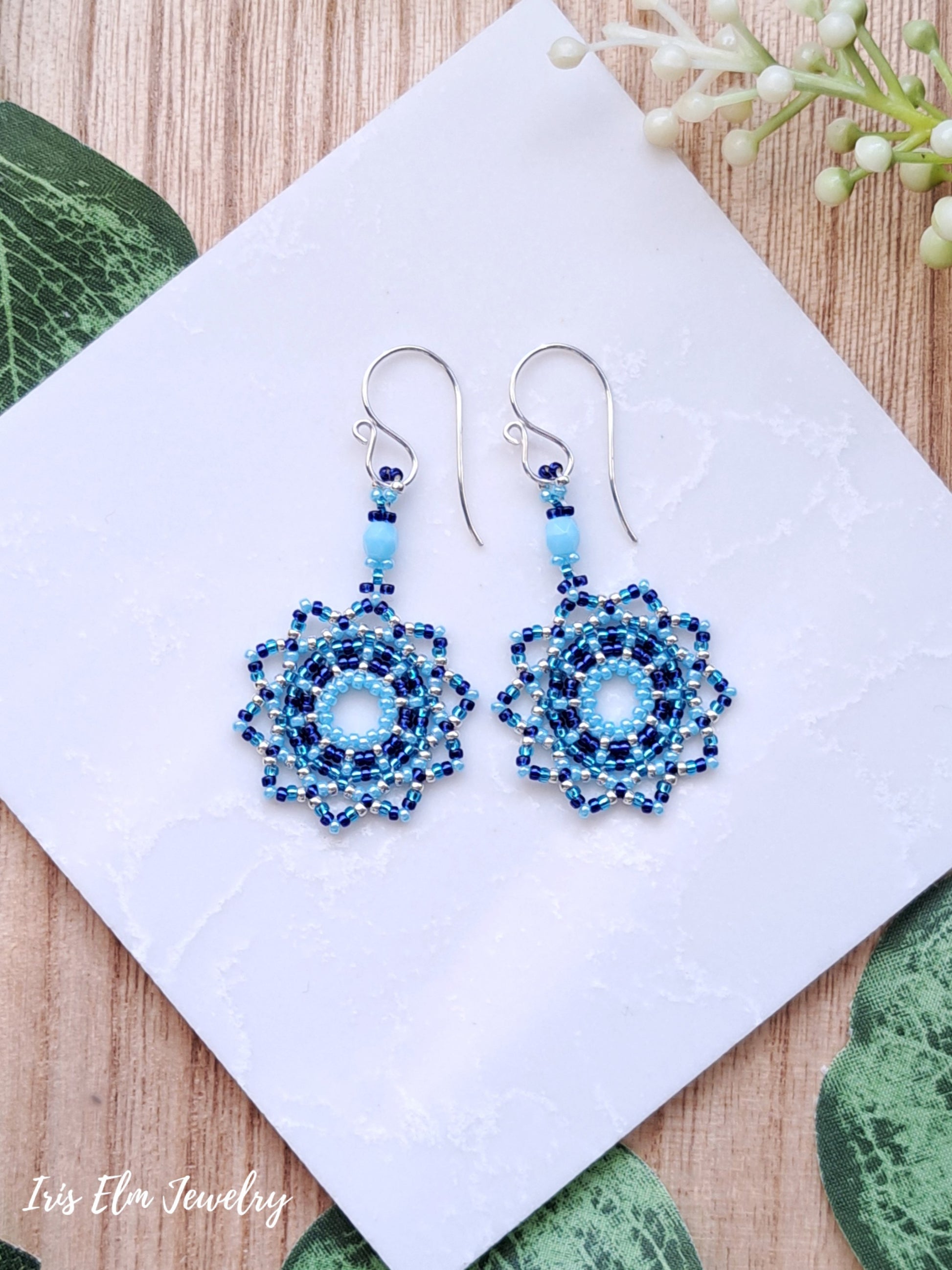 Blue beaded seed bead mandala earrings on a white card with green leaves and flowers in the background