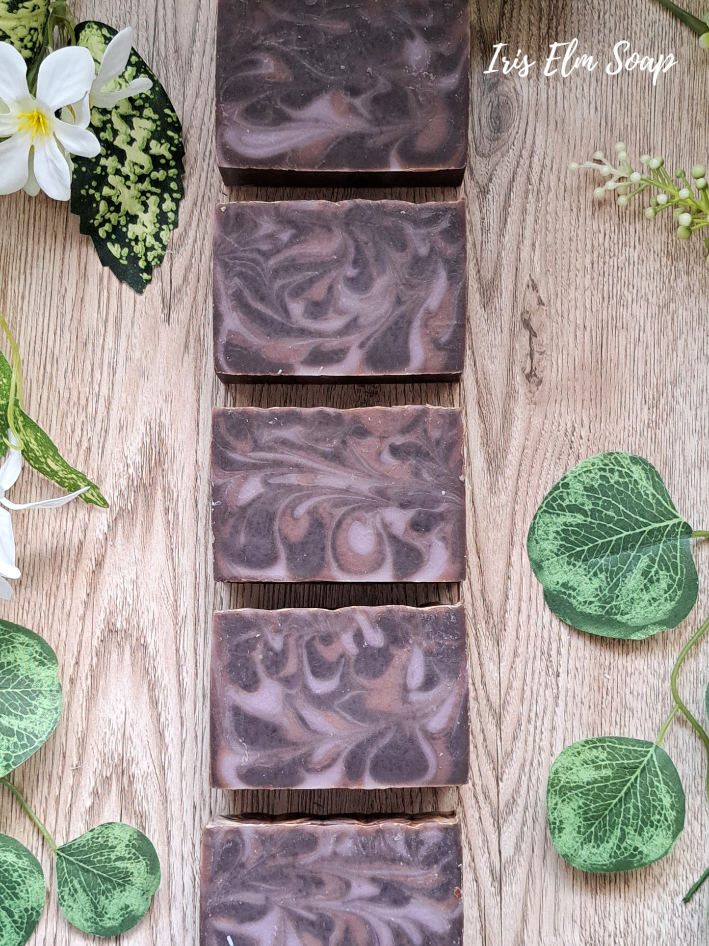 Marbled purple and brown black amber and lavender handmade soap bars on a wooden surface with green leaves and flowers.