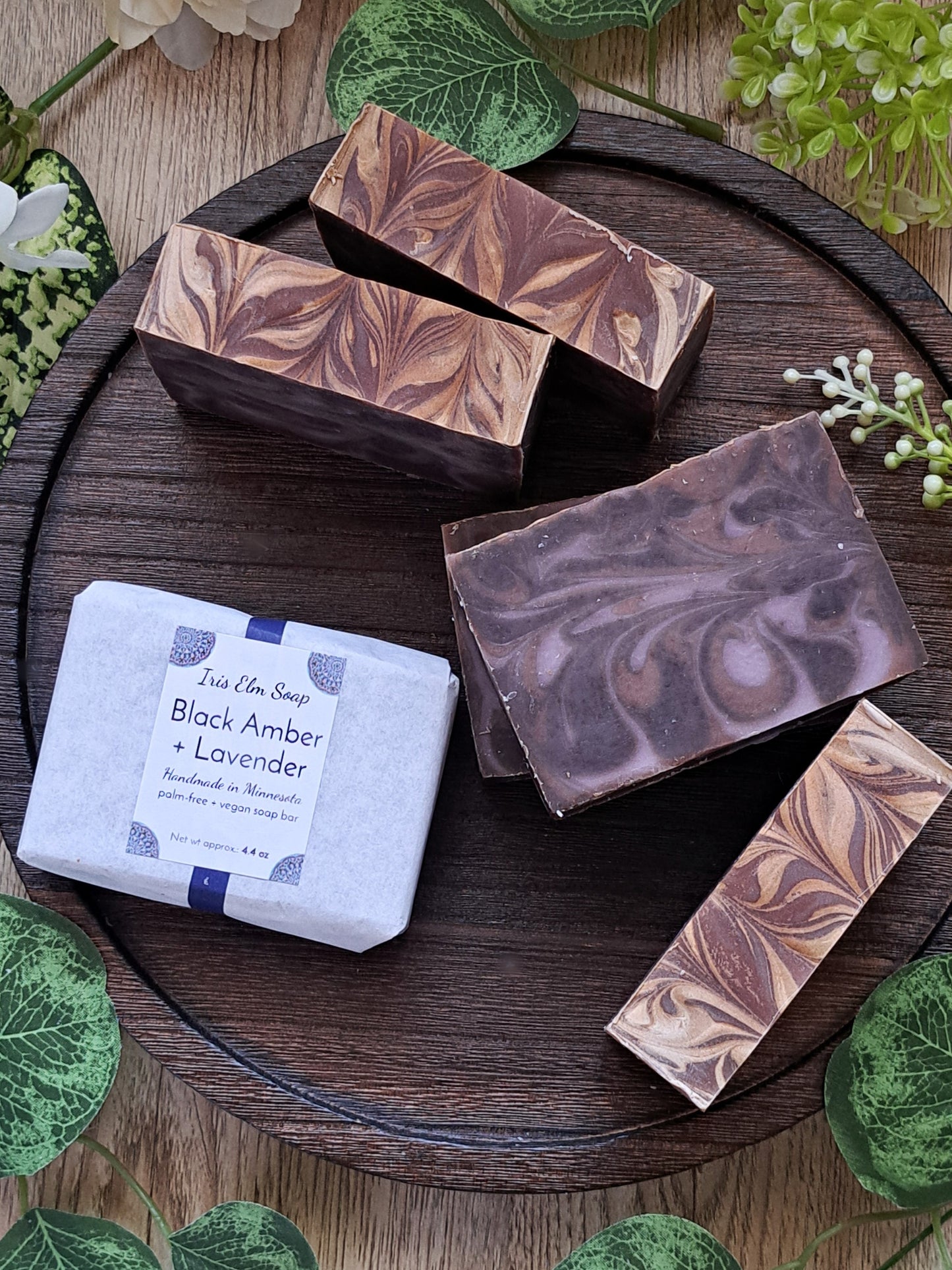 Marbled black amber and lavender handmade soap bars on a wooden tray with greenery around