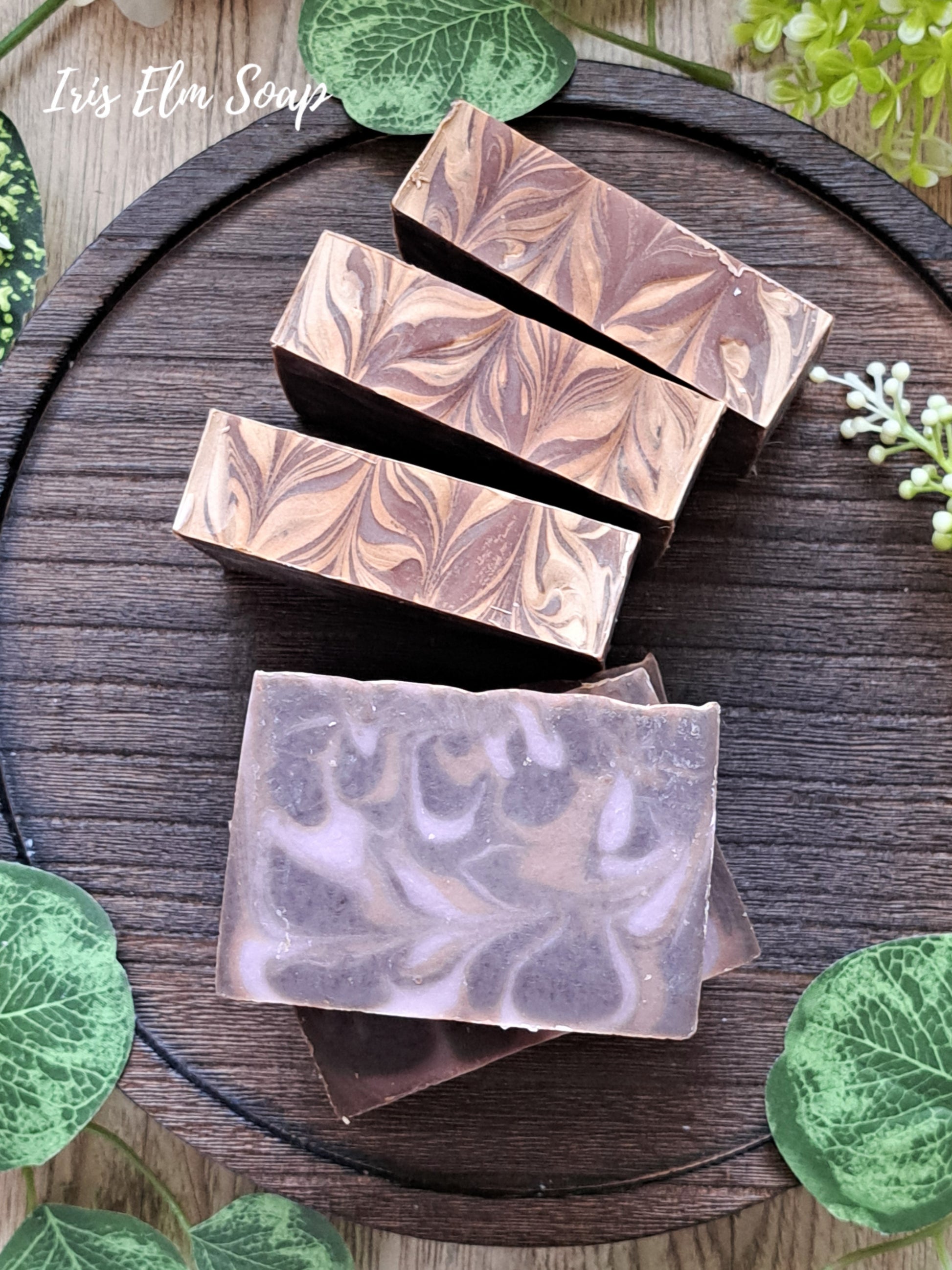 Five bars of black amber and lavender handmade soap with a marbled design on a wooden surface, surrounded by green leaves.