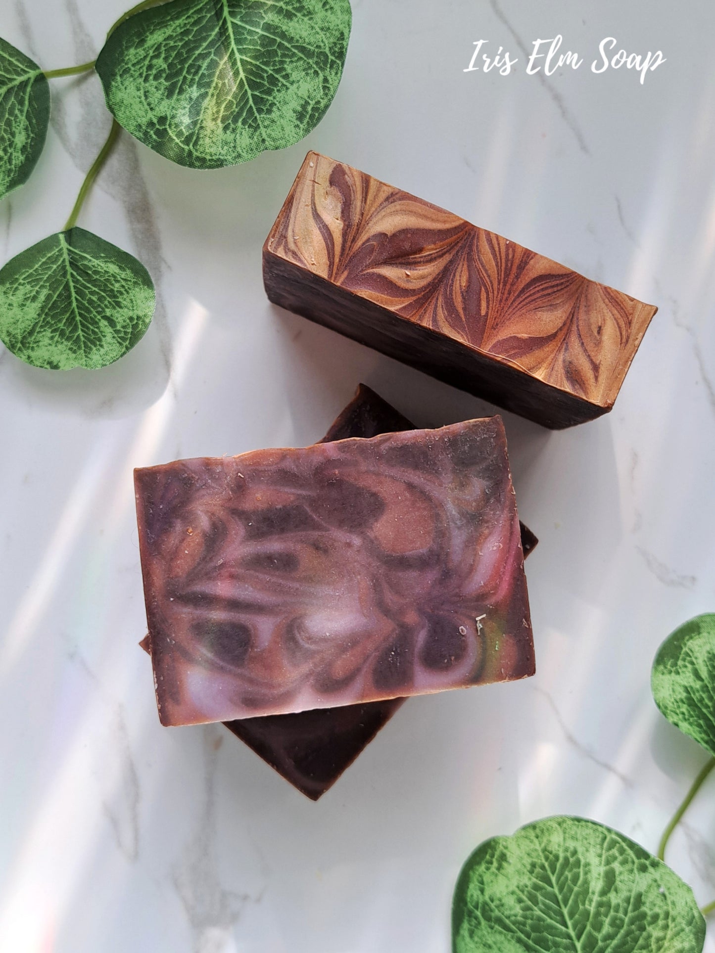 Two brown and purple marbled black amber and lavender handmade soap bars on a white surface with green leaves.