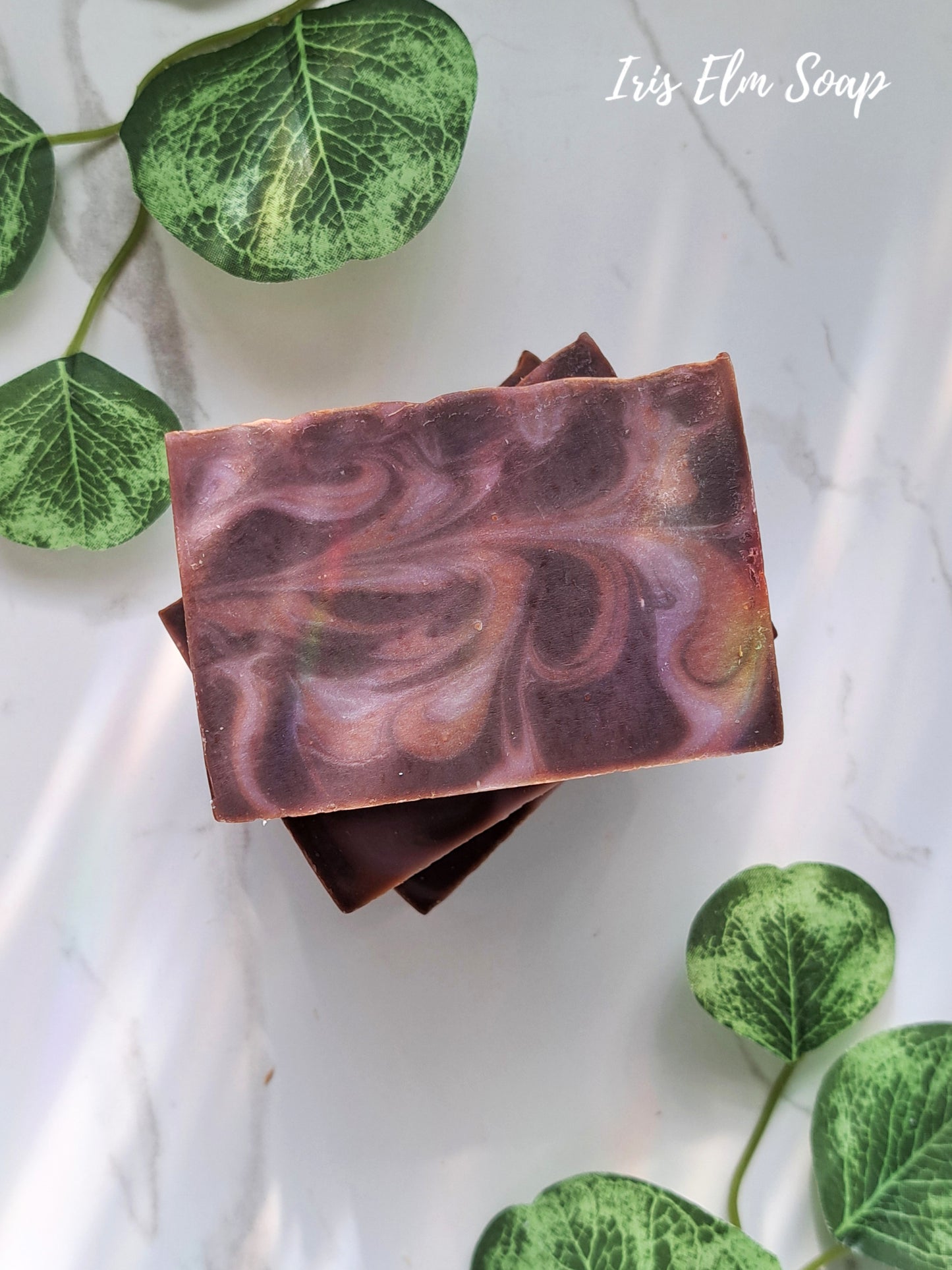 Two swirled purple and brown black amber and lavender handmade soap bars on a white background with green leaves.