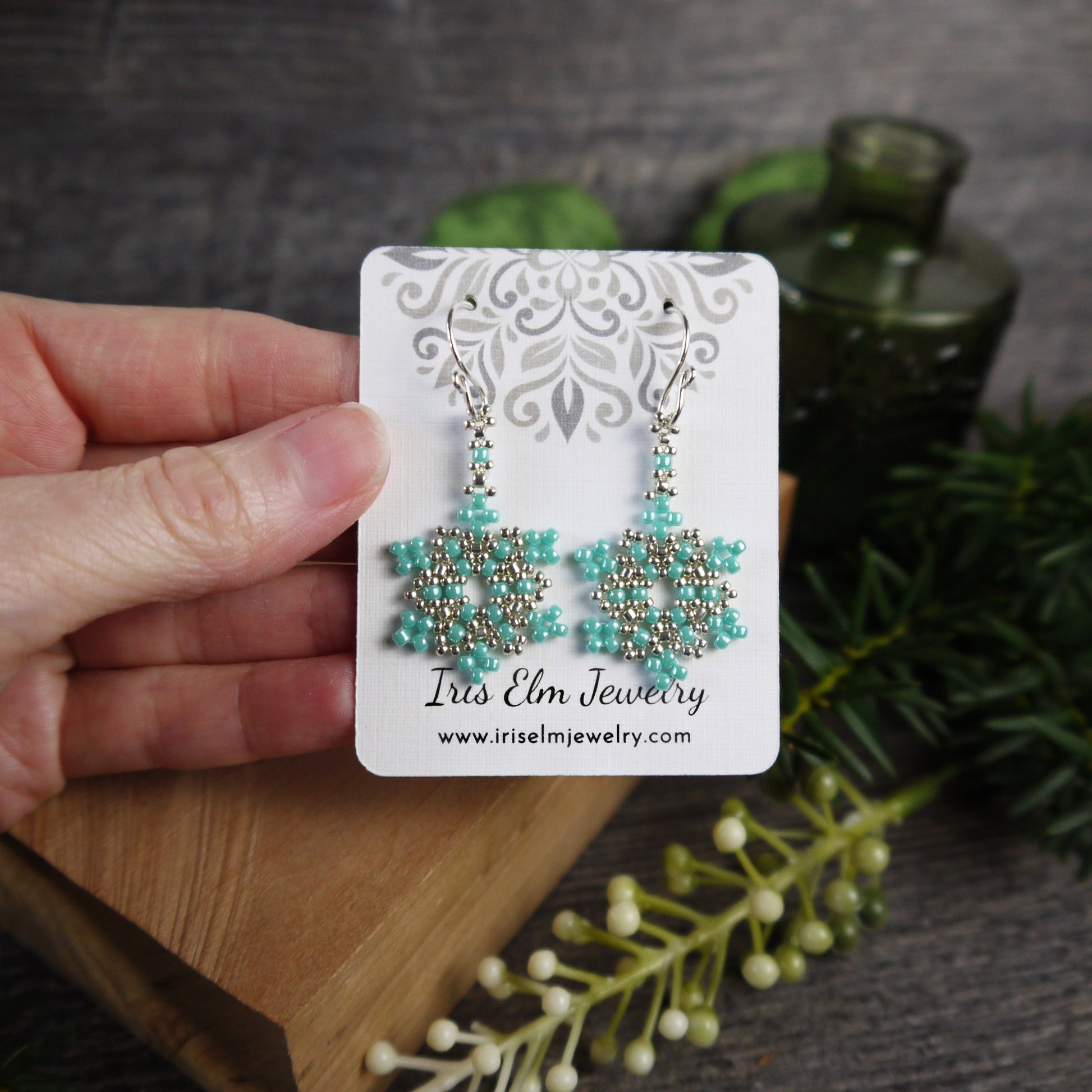 Turquoise and silver beaded glass snowflake earrings on a 'Iris Elm Jewelry' card held by a hand with a natural background.