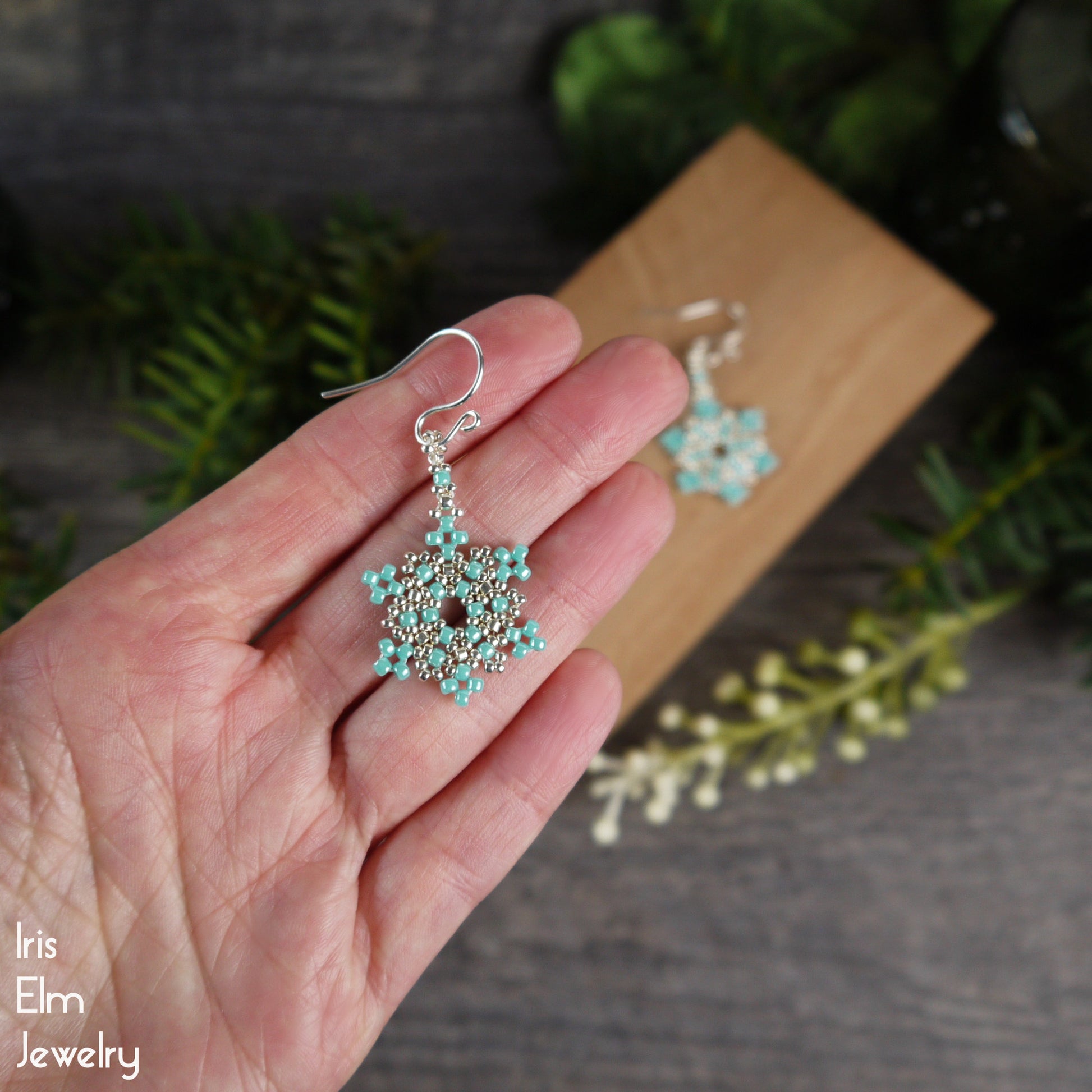 Hand holding a turquoise glass seed bead woven snowflake beaded earring with 'Iris Elm Jewelry' branding.