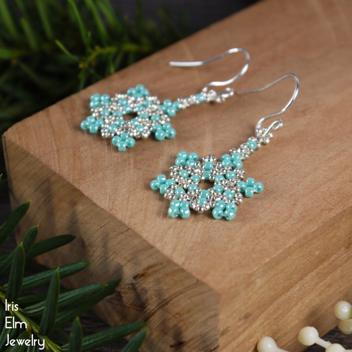 Turquoise glass seed bead beaded snowflake earrings on a wooden block with 'Iris Elm Jewelry' branding.