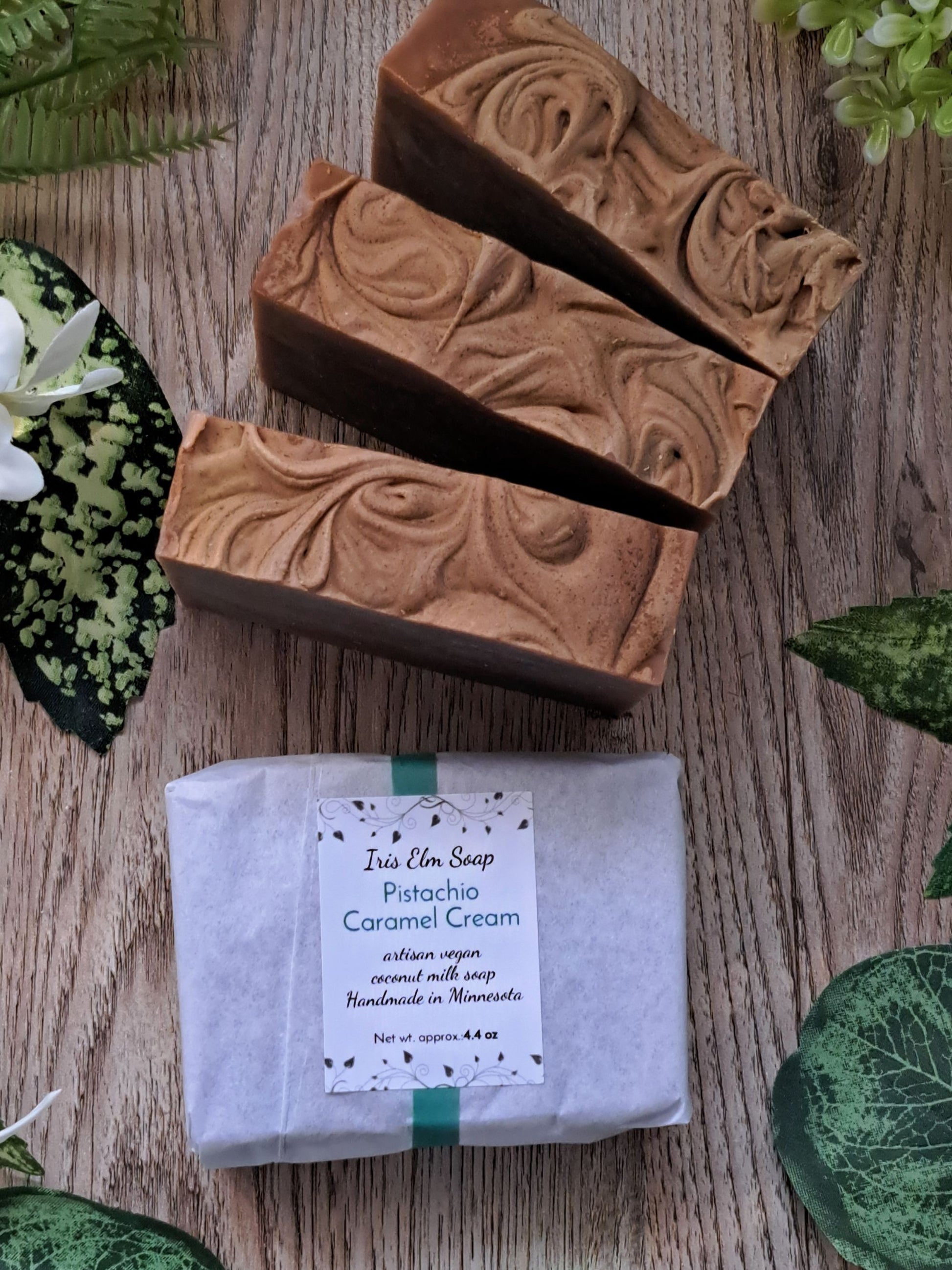 Three bars of pistachio caramel cream handmade soap with a wrapped bar on a wooden surface with greenery.