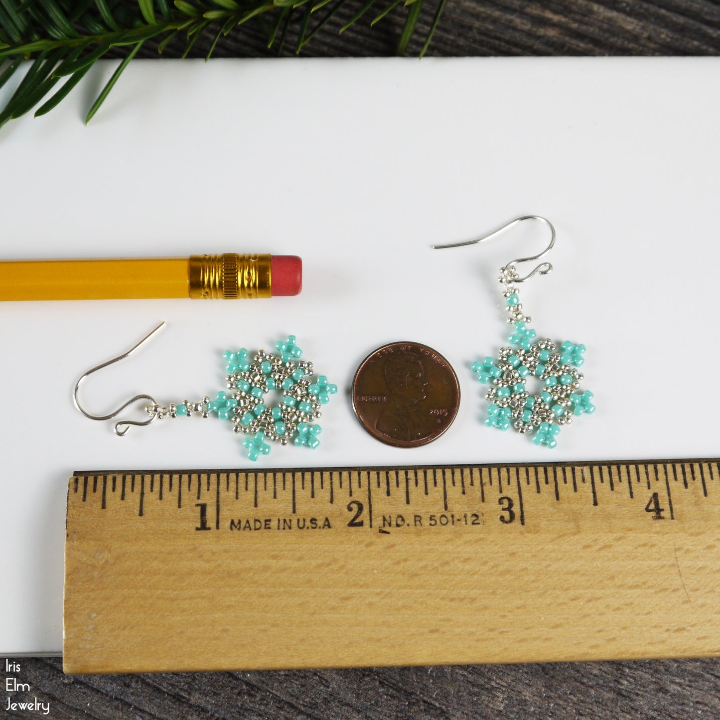 Pair of 1.75 inch long turquoise seed bead snowflake beaded earrings on a white surface with a ruler for scale.