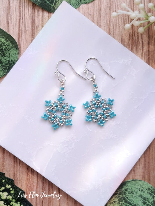 Pair of turquoise beaded seed bead snowflake earrings on a white napkin with 'Iris Elm Jewelry' branding.