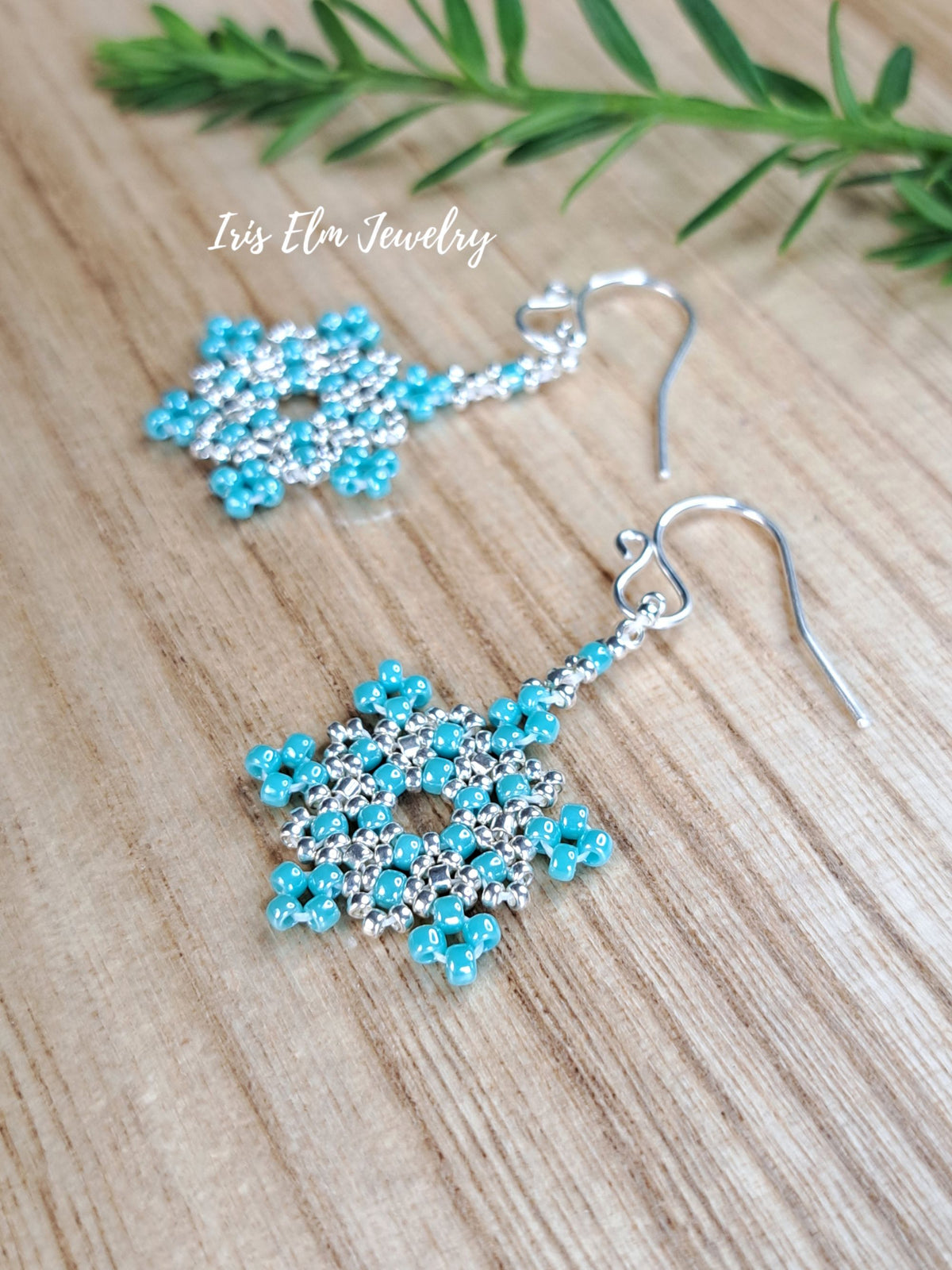 A pair of handmade snowflake-shaped dangle earrings made with turquoise and silver seed beads, displayed on a light wood surface with a green sprig in the background. The brand name 'Iris Elm Jewelry' is watermarked on the image.