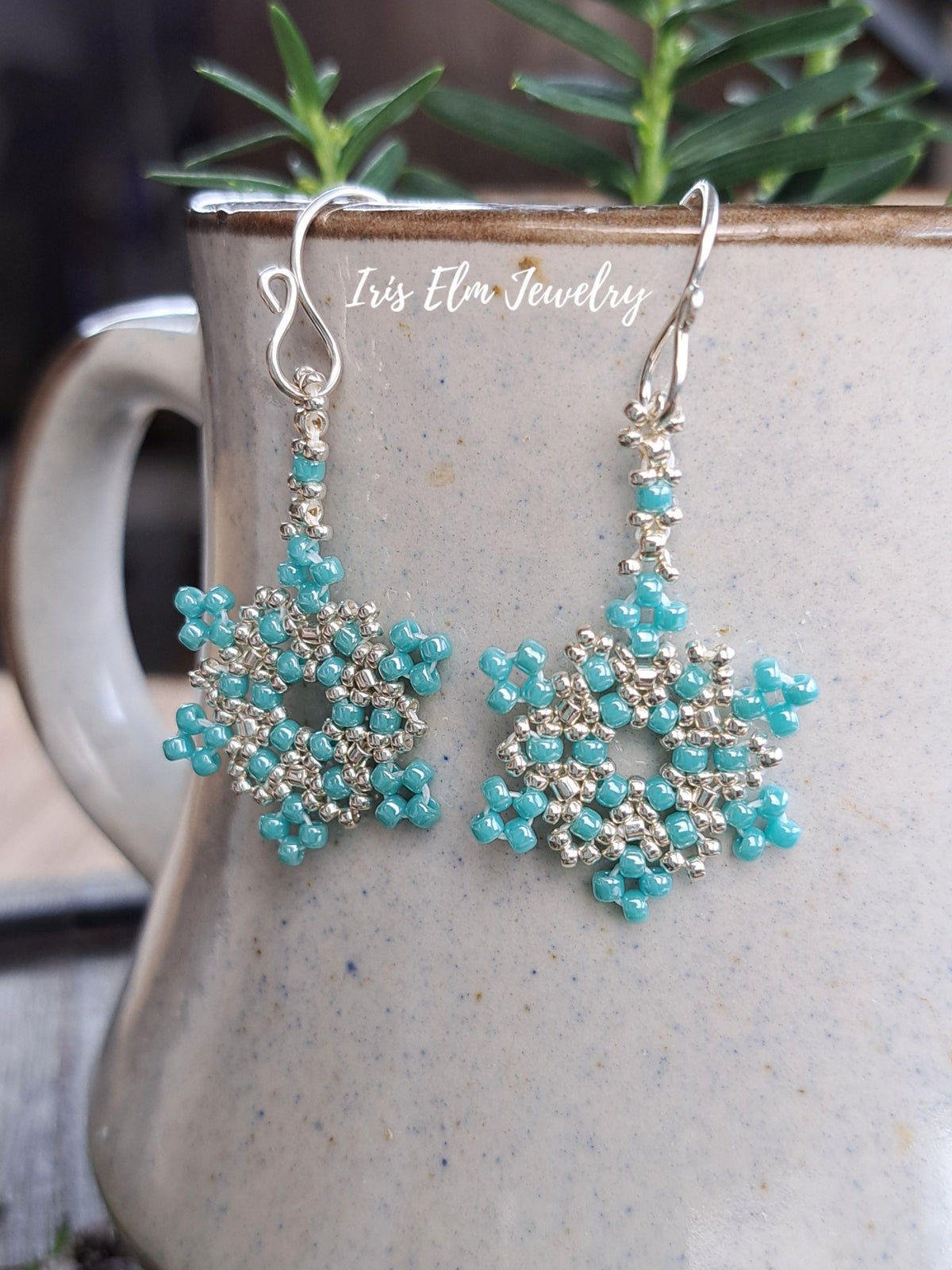 A pair of handmade beaded dangle earrings with turquoise blue and silver seed beads forming snowflake-like patterns, hanging from silver fishhook wires. The earrings are displayed against a speckled beige ceramic mug with green foliage in the background.
