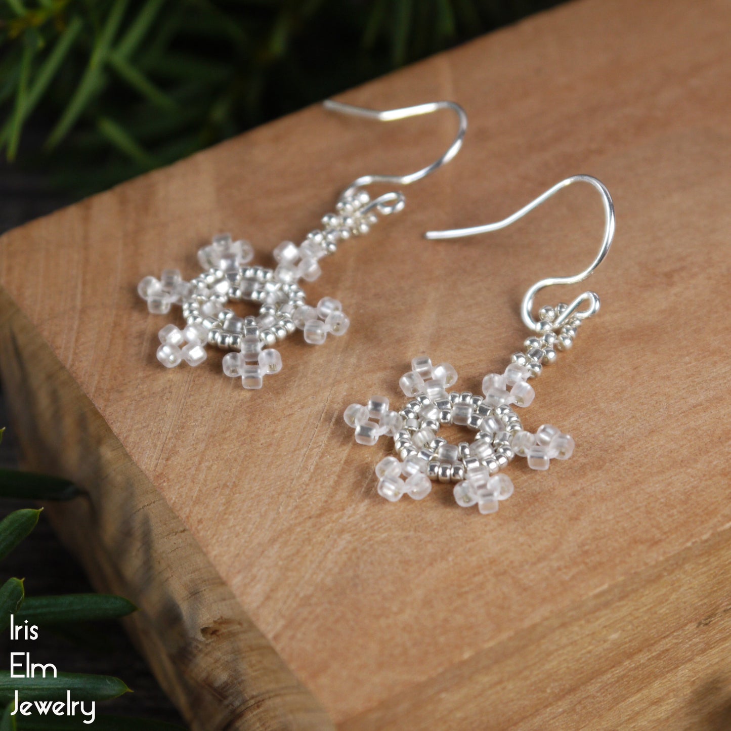 Silver and white seed bead snowflake earrings on a wooden surface with 'Iris Elm Jewelry' branding.