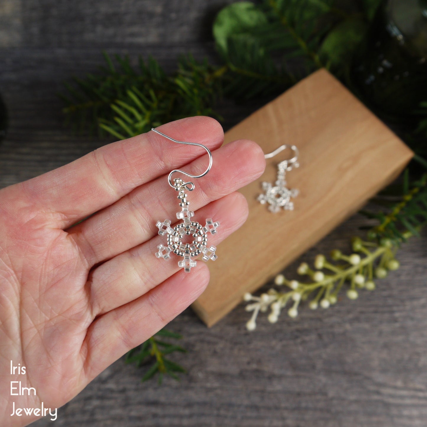 White beaded snowflake earrings held in a hand with a wooden block and greenery in the background.