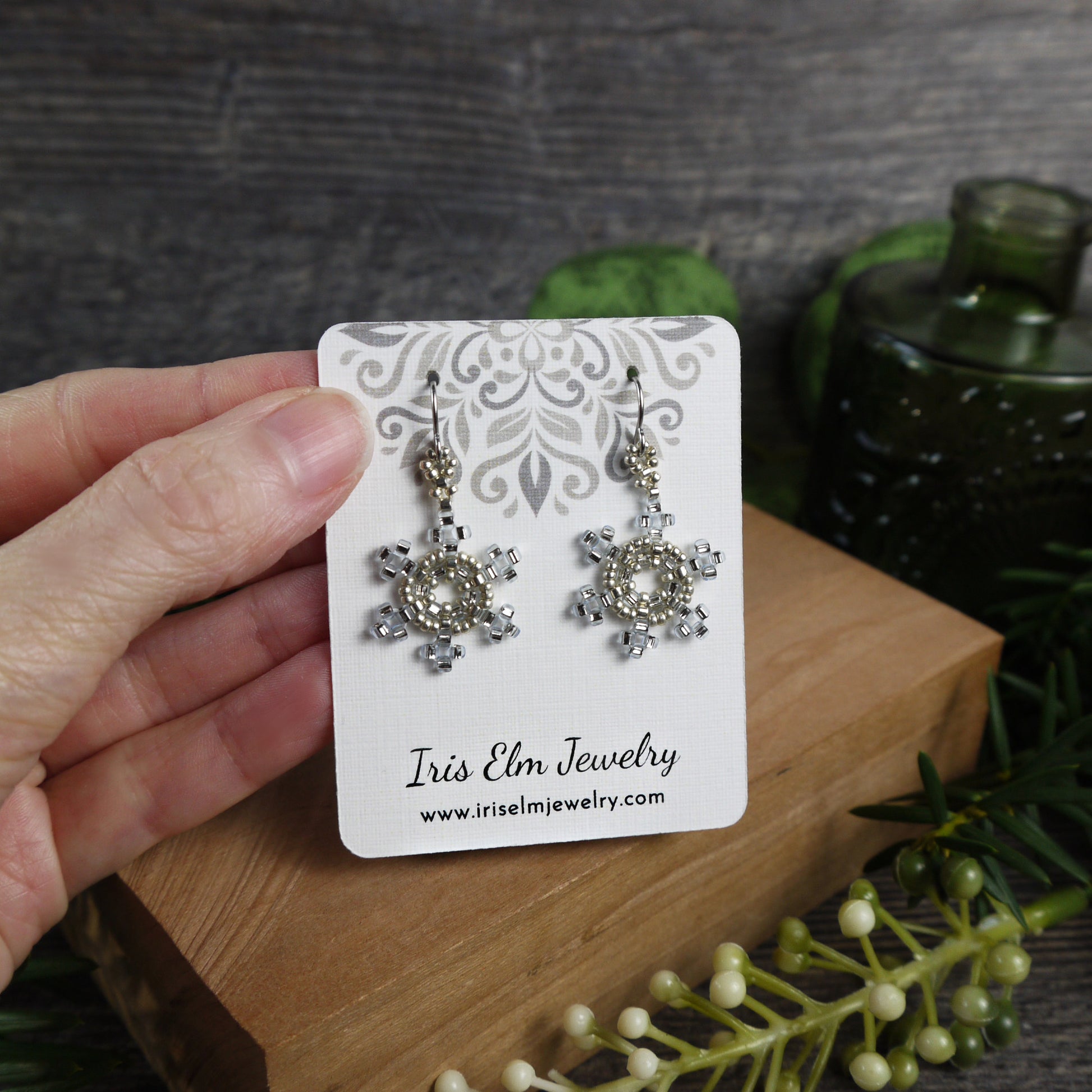 Pair of snowflake-shaped earrings on a 'Iris Elm Jewelry' card held by a hand with a natural background.