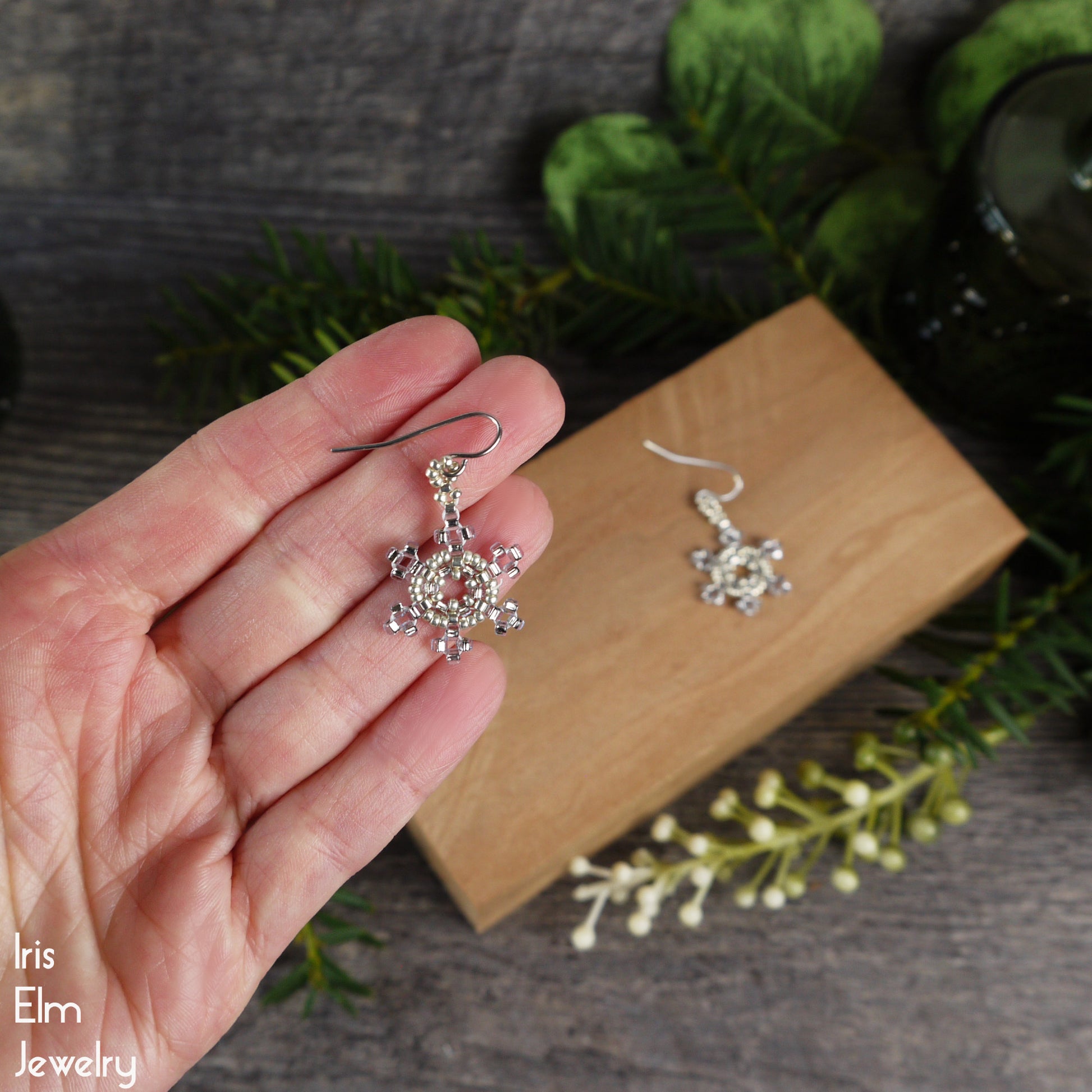 Silver and pale blue glass seed bead snowflake earrings held in a hand with a wooden box and greenery in the background.