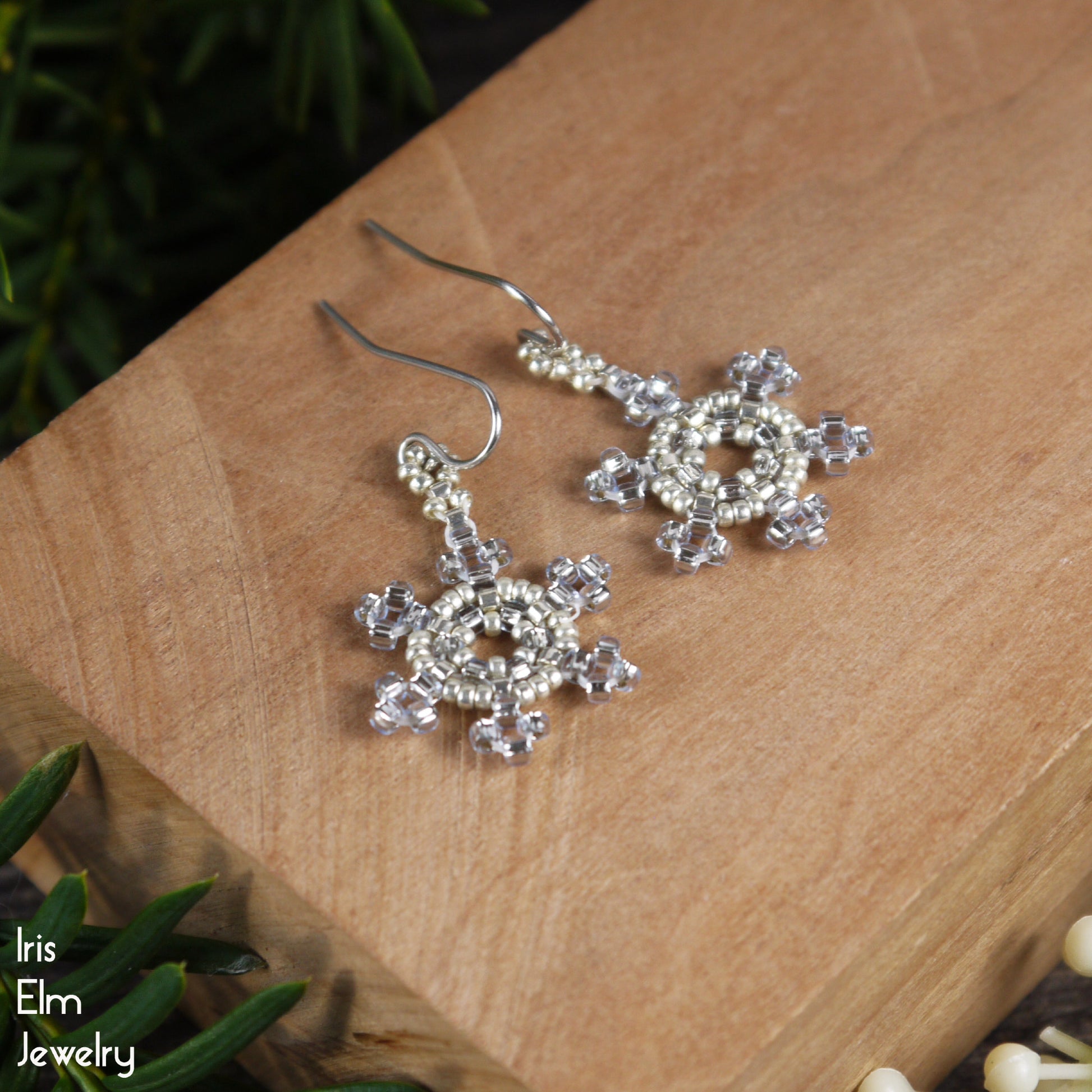 Silver and light icy blue seed bead woven snowflake earrings on a wooden surface with 'Iris Elm Jewelry' branding.