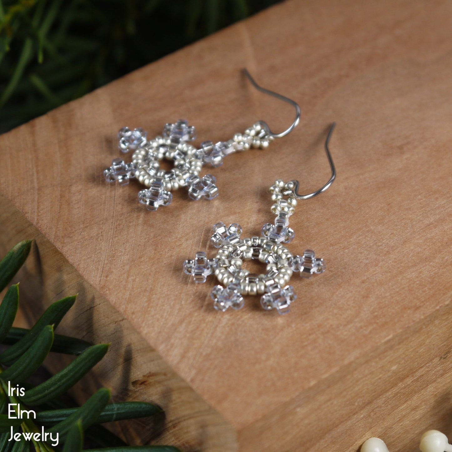 Silver and light blue glass seed bead handmade snowflake earrings on a wooden surface with greenery