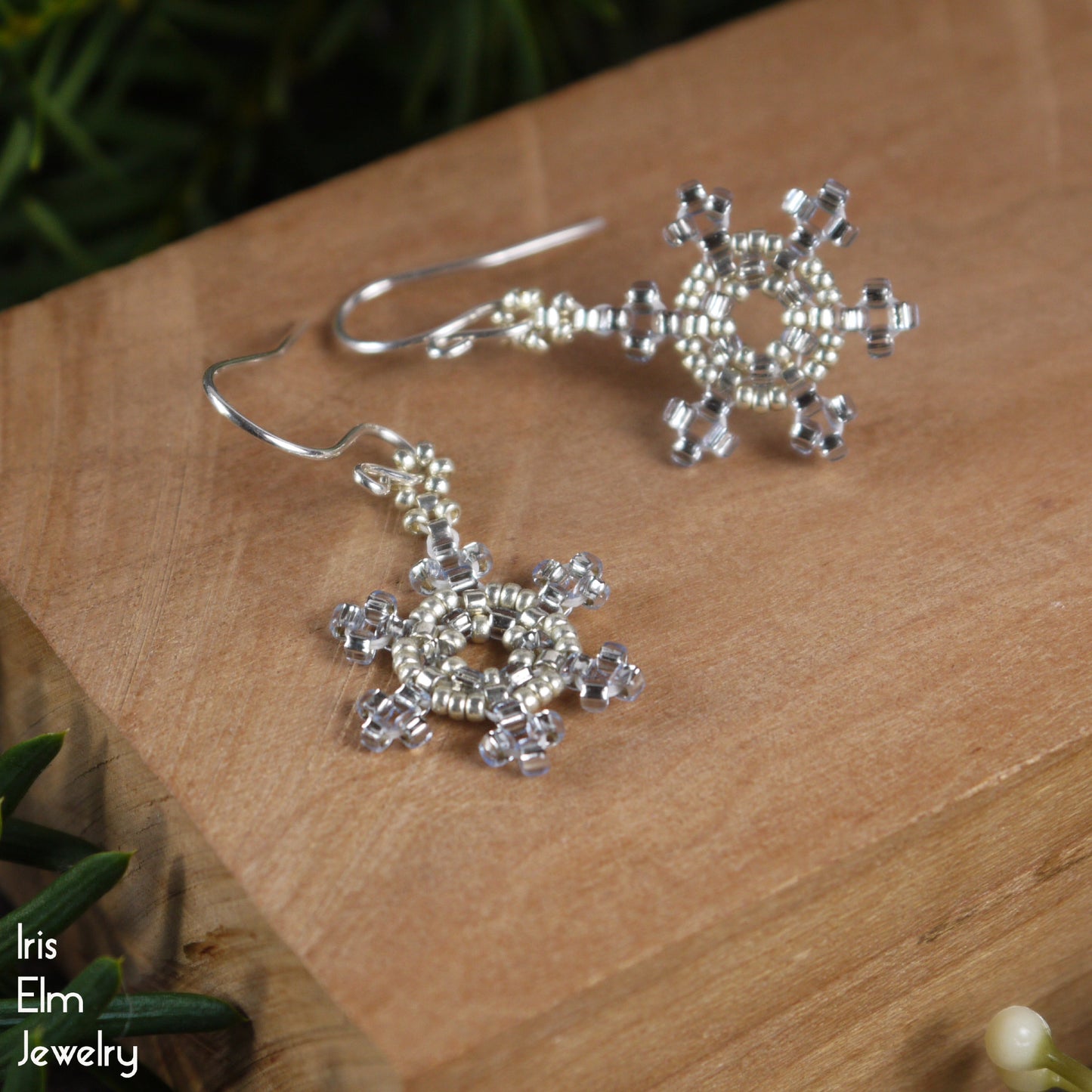 Silver and pale blue seed bead snowflake earrings on a wooden surface with 'Iris Elm Jewelry' branding.