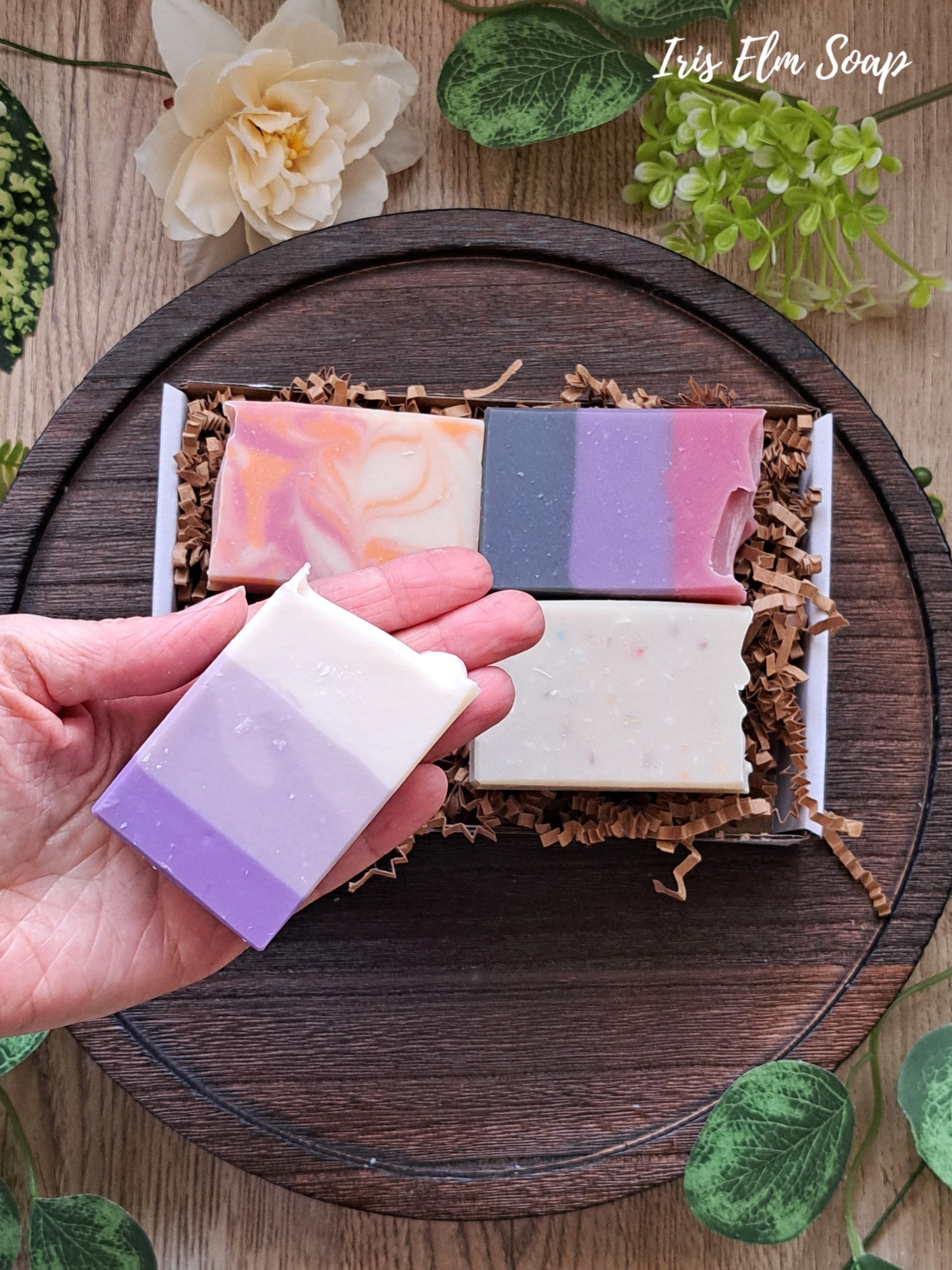 Hand holding a striped soap bar with more soap bars in gift box in the background on a wooden surface.