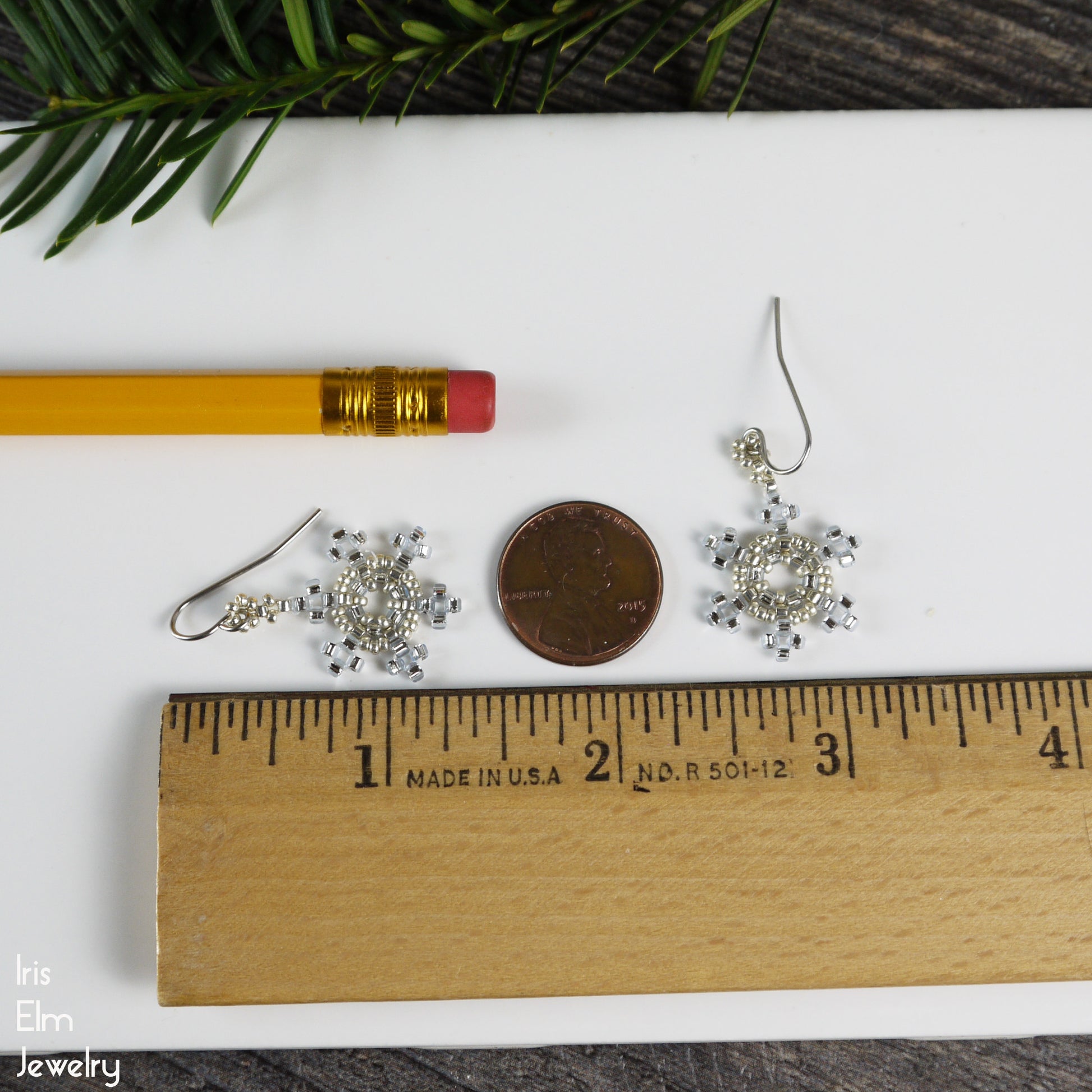 Silver 1 and 3/8 inch long snowflake earrings on a white surface with a pencil, coin, and ruler for scale.