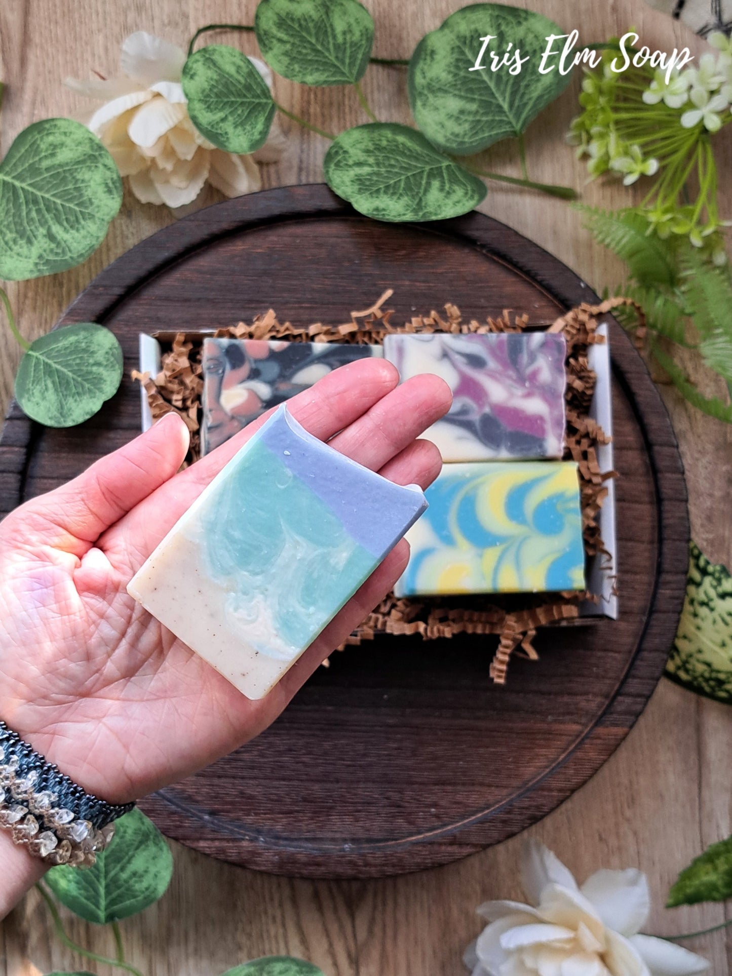 Hand holding a bar of soap with other bars on a wooden tray, surrounded by greenery and flowers.